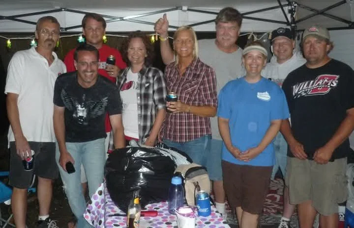 Group of nine people standing together outdoors under a white canopy tent, smiling and posing for a photo. There is a table with drinks and a black trash bag in front of them.