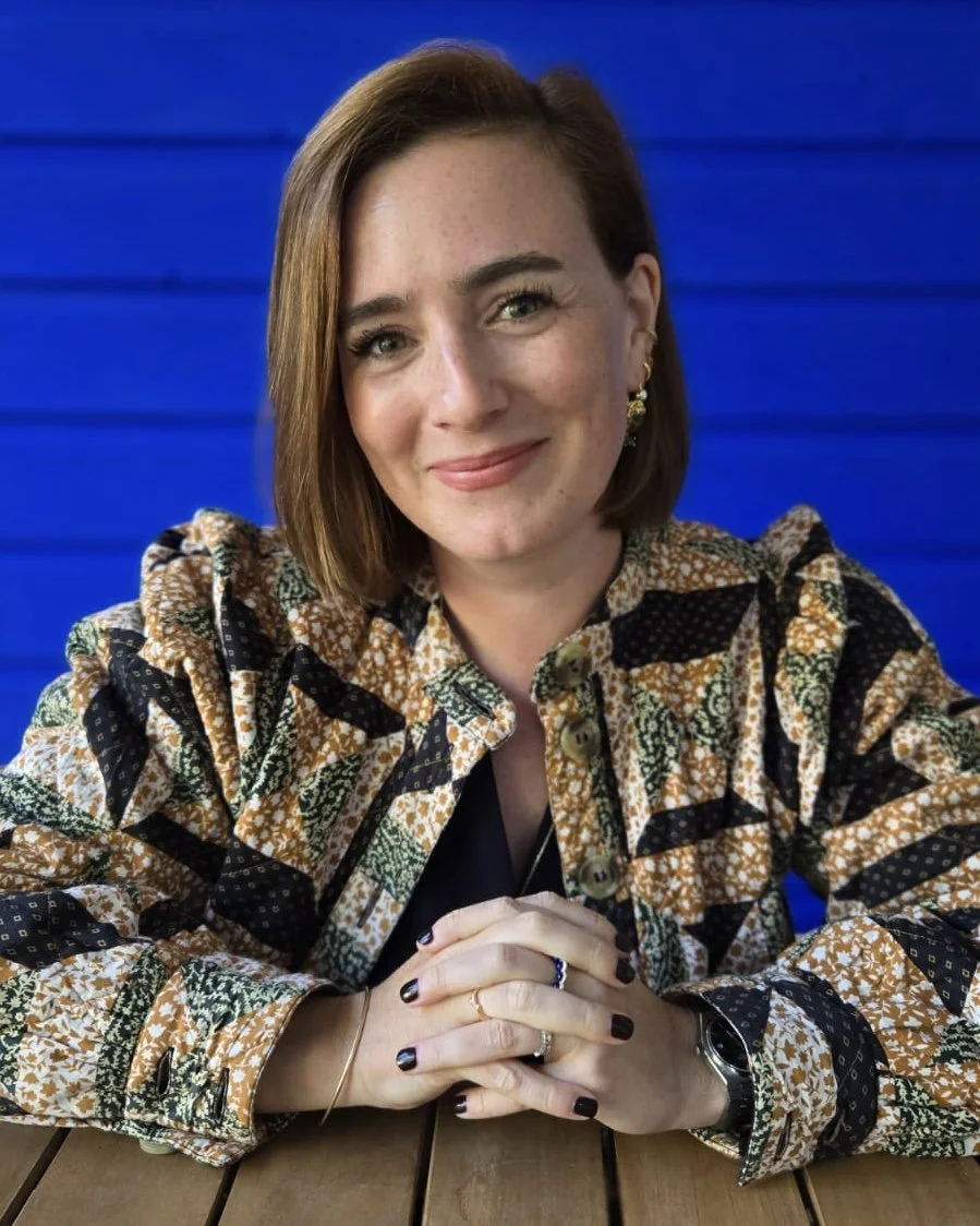 Une femme aux cheveux courts, portant une chemise à motifs colorés, souriant, assise à une table en bois devant un fond bleu.