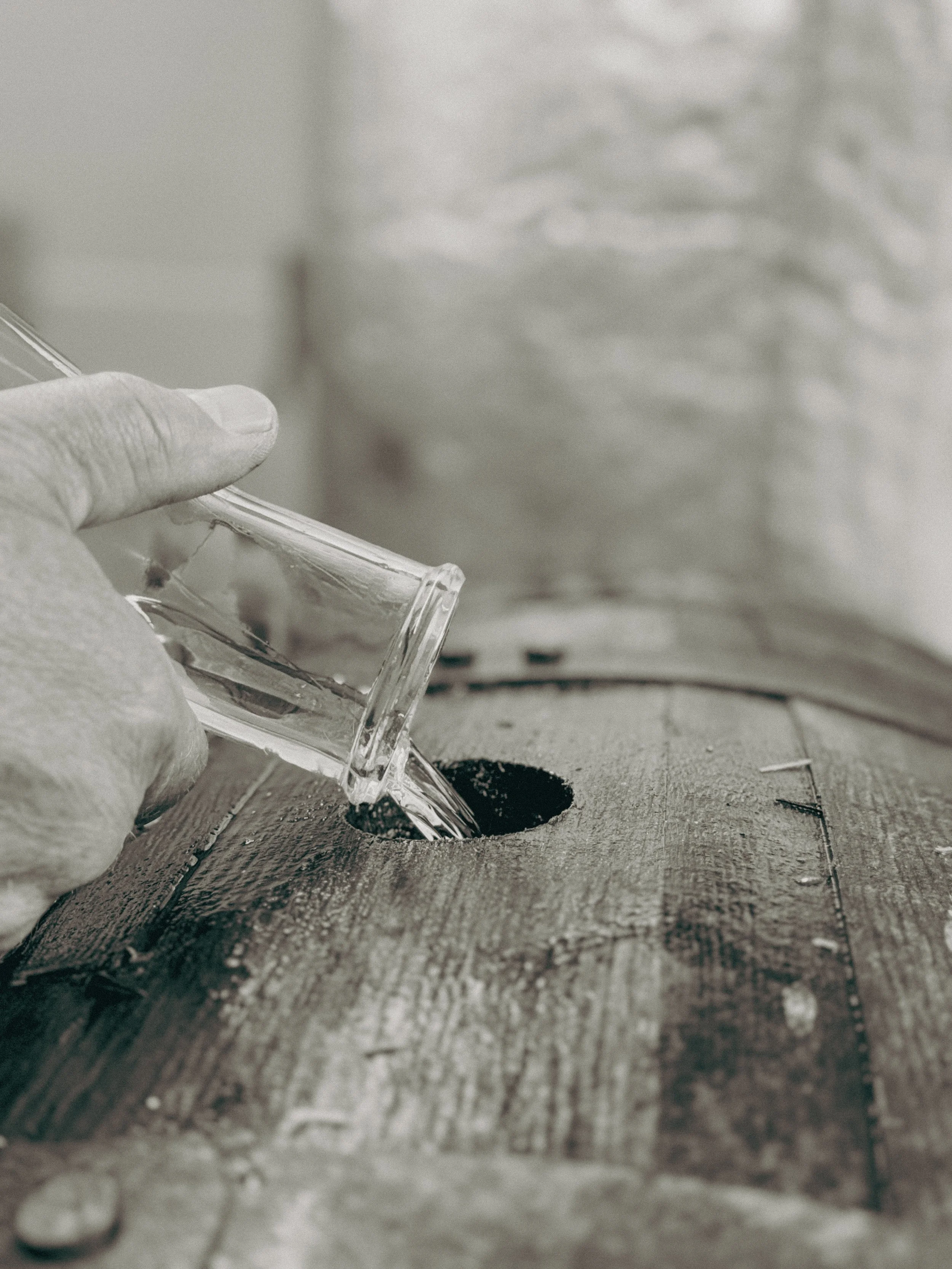 Blackhouse Rum, BH monogram logo, Off Grid distillery, Glenuig, Moidart, Craft rum, Scottish Rum, Craft Spirit, Small batch RumA hand holding a glass container pouring liquid into a hole in a wooden surface.