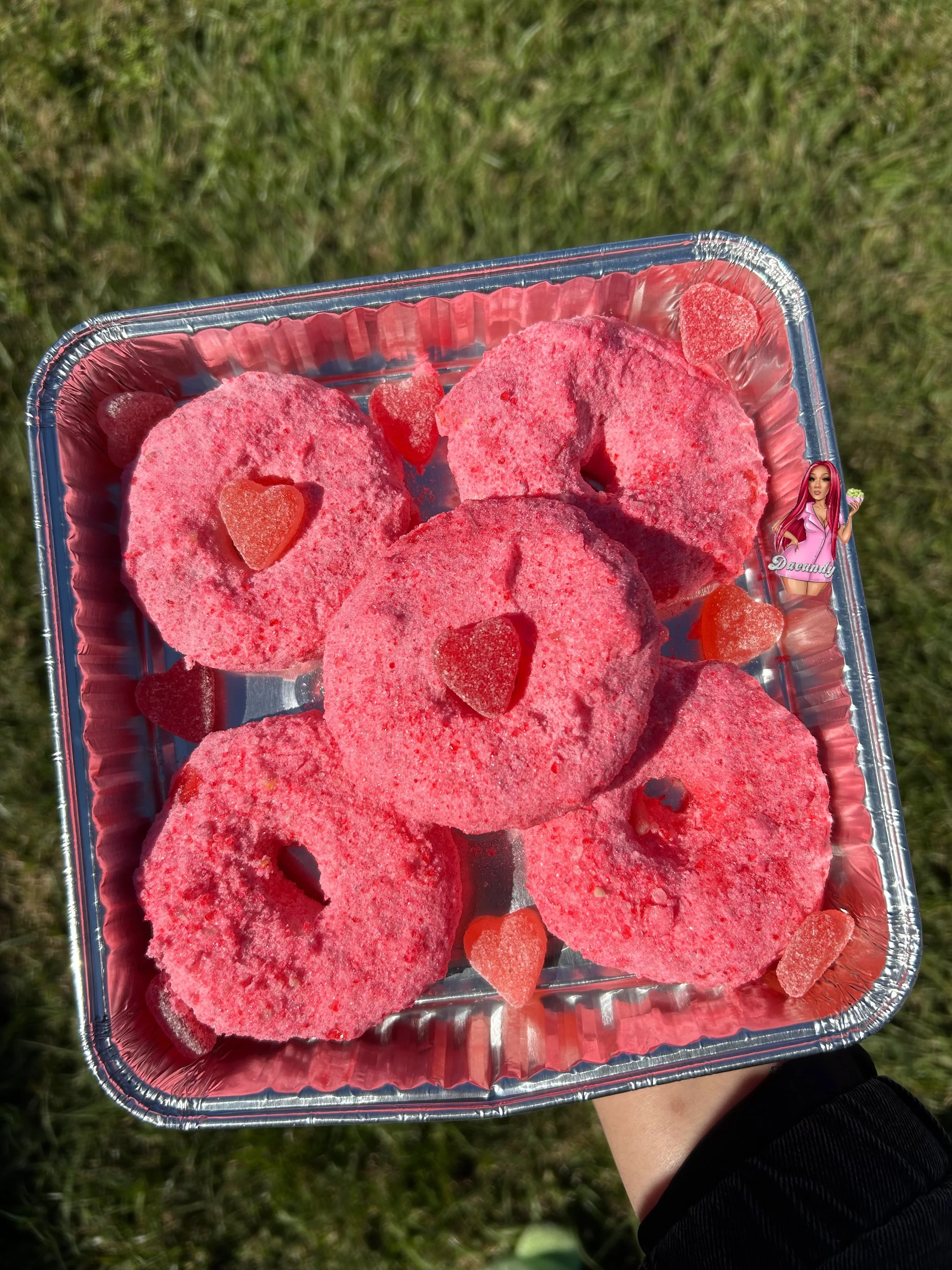 Pink frosted donuts with red heart-shaped gummy candies on top and around in a disposable aluminum tray, held outdoors over grass.