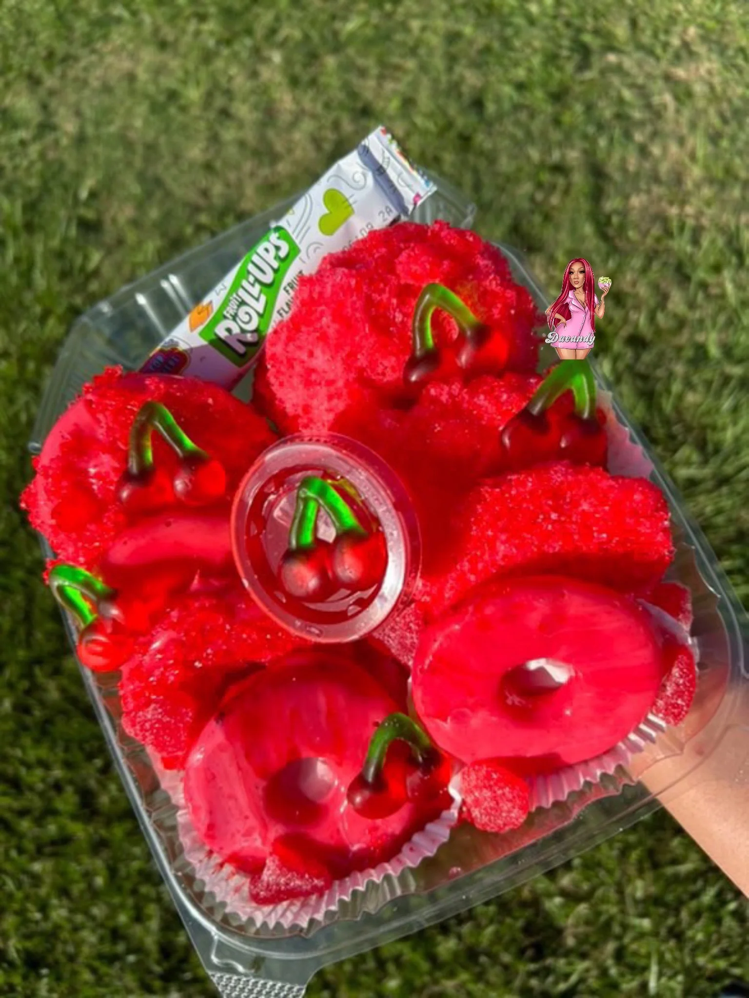 A hand holding a clear plastic container with red cherry-shaped desserts and a packet of Rainbow Roll candies, set against a background of grass.