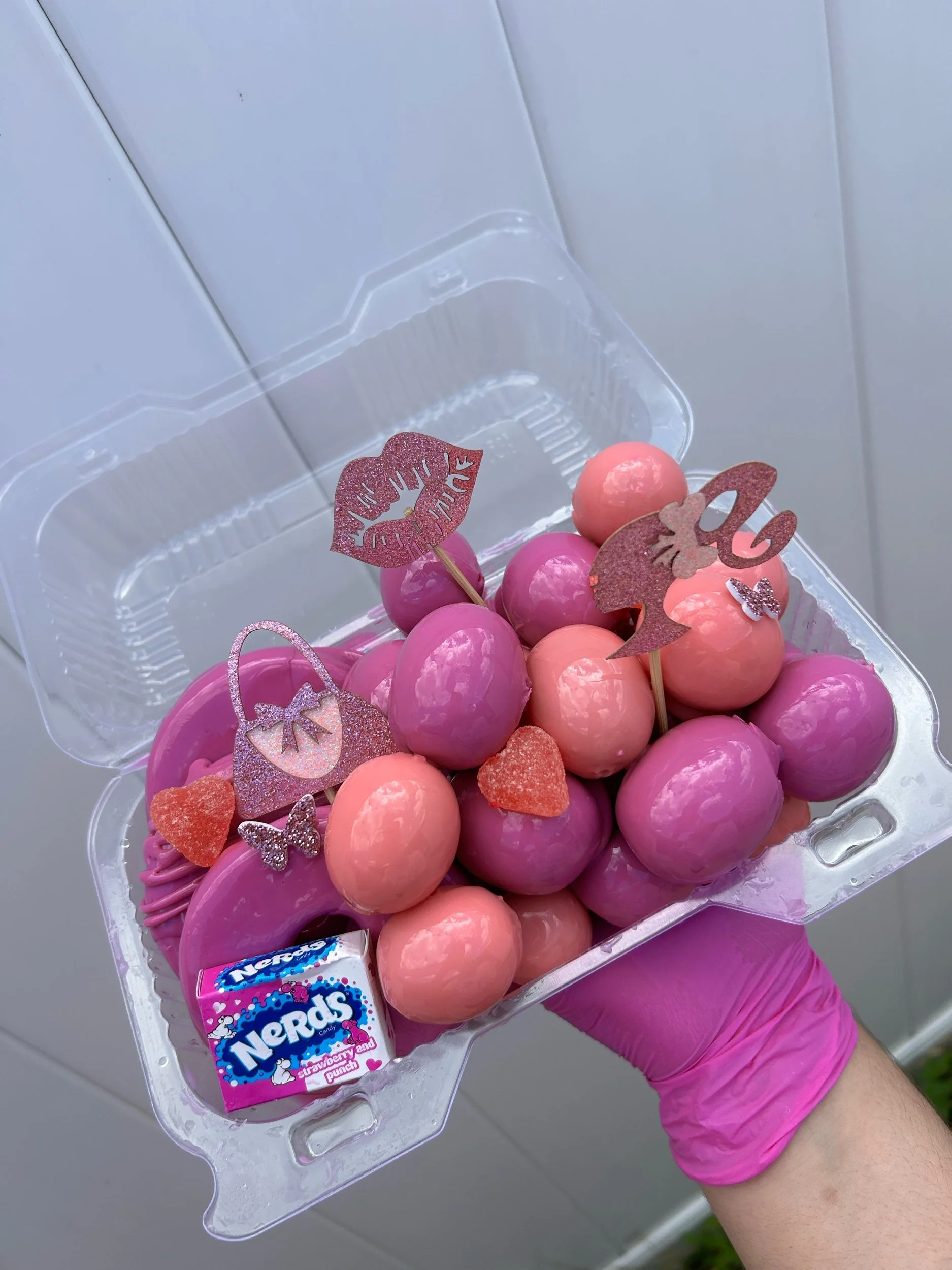 Pink, purple, and peach cake pops decorated with glittery paper and candy shapes, along with a small box of Nerds candy, displayed in a clear plastic container.