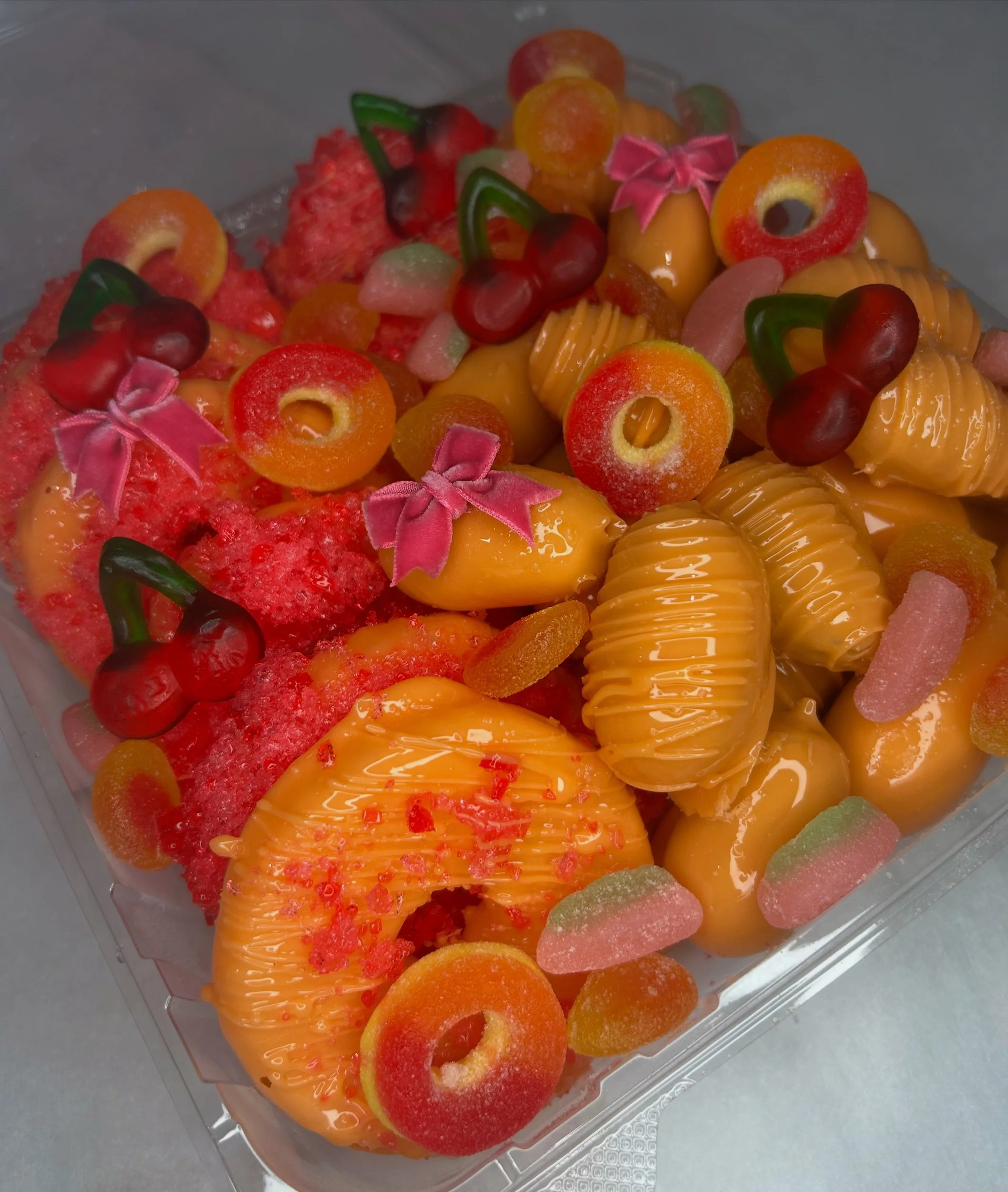 A colorful display of assorted candies and gummy treats, including peach rings, gummy cherries, and other fruit-shaped gummies, in a clear plastic container.