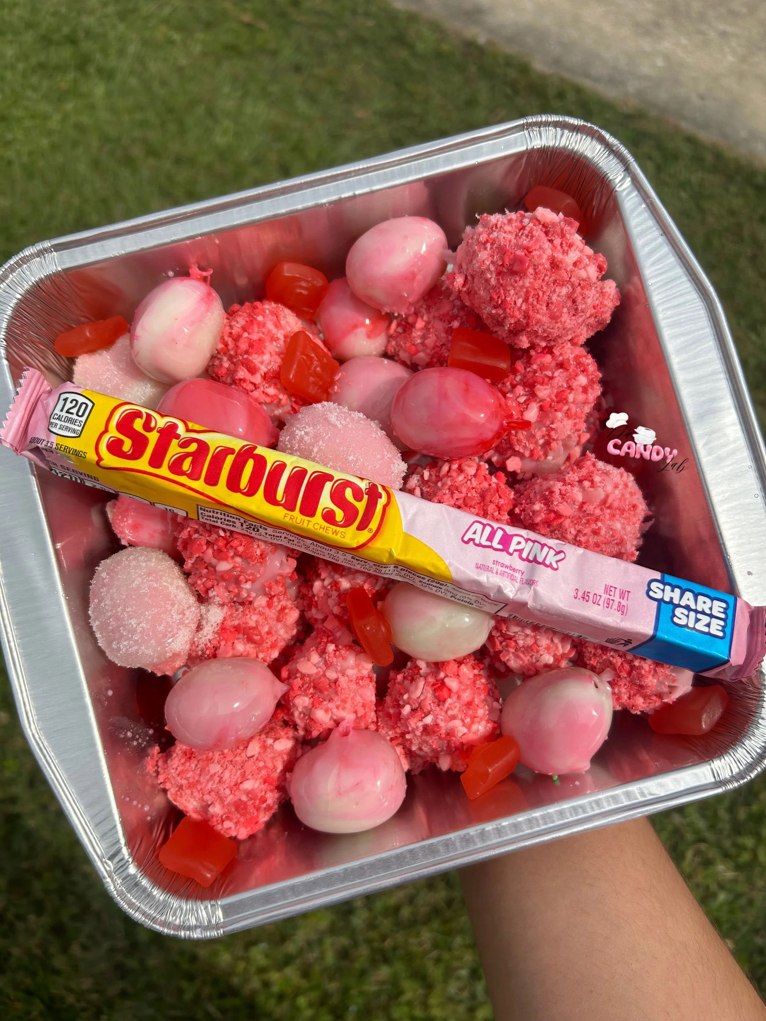 A metal tray filled with pink and white candies, including a Starburst fruit chew, cherry-flavored gummies, and sugared gummy balls, held outdoors over grass.
