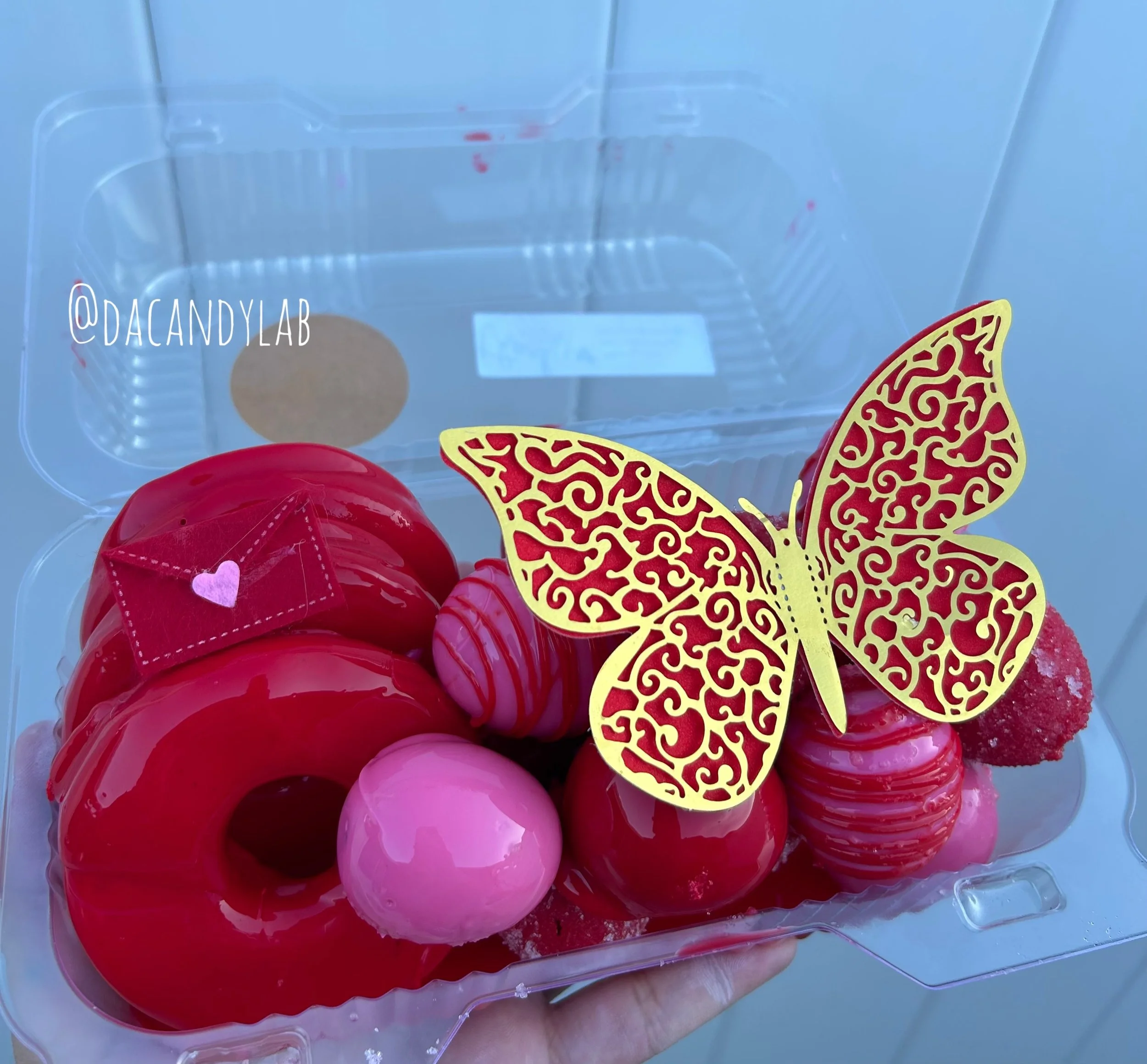 A clear plastic container holding an assortment of pink and red domed donuts with a decorative yellow and red butterfly ornament on top.