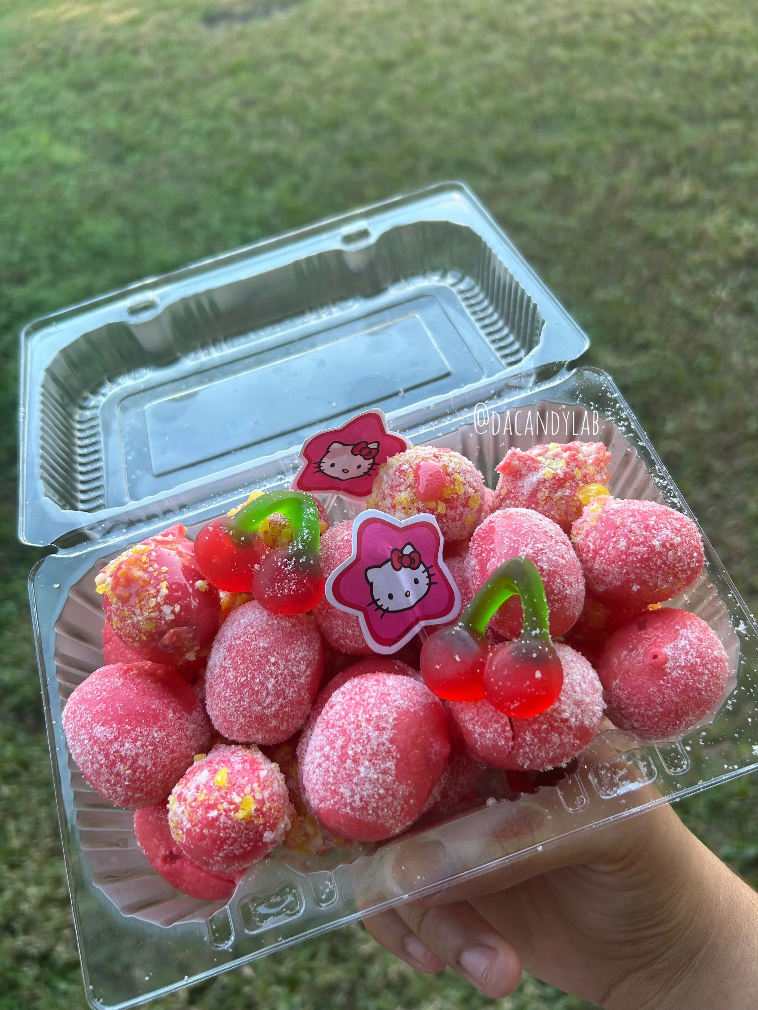 A clear plastic container holds pink candy strawberries decorated with powdered sugar and topped with cherry-shaped gummy candies. The container has Hello Kitty-themed stickers on the lid.