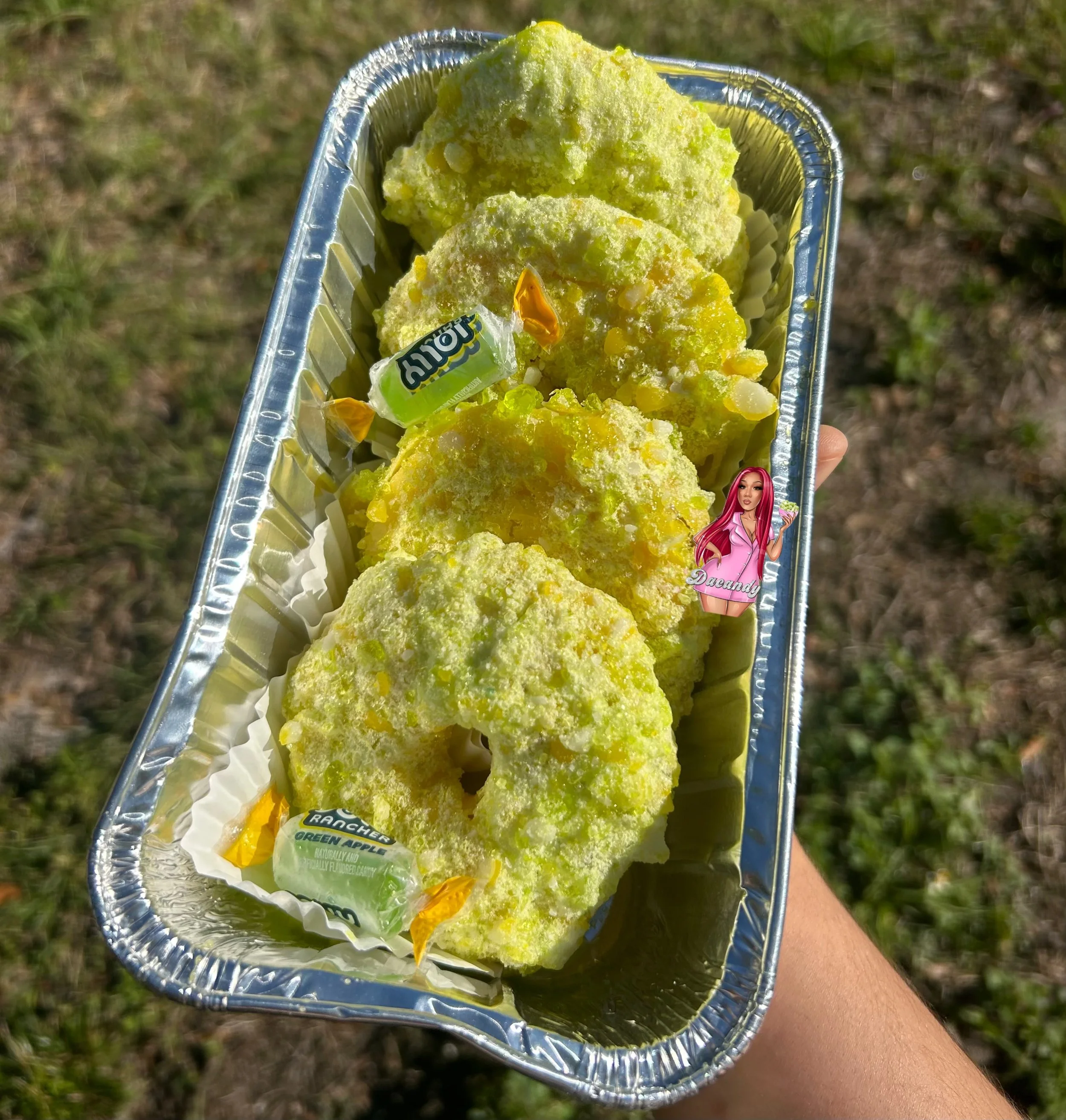 Three slices of green apple-flavored sno cone in a silver foil tray, with a piece of bubble gum and a sticker of a woman with pink hair on the side.