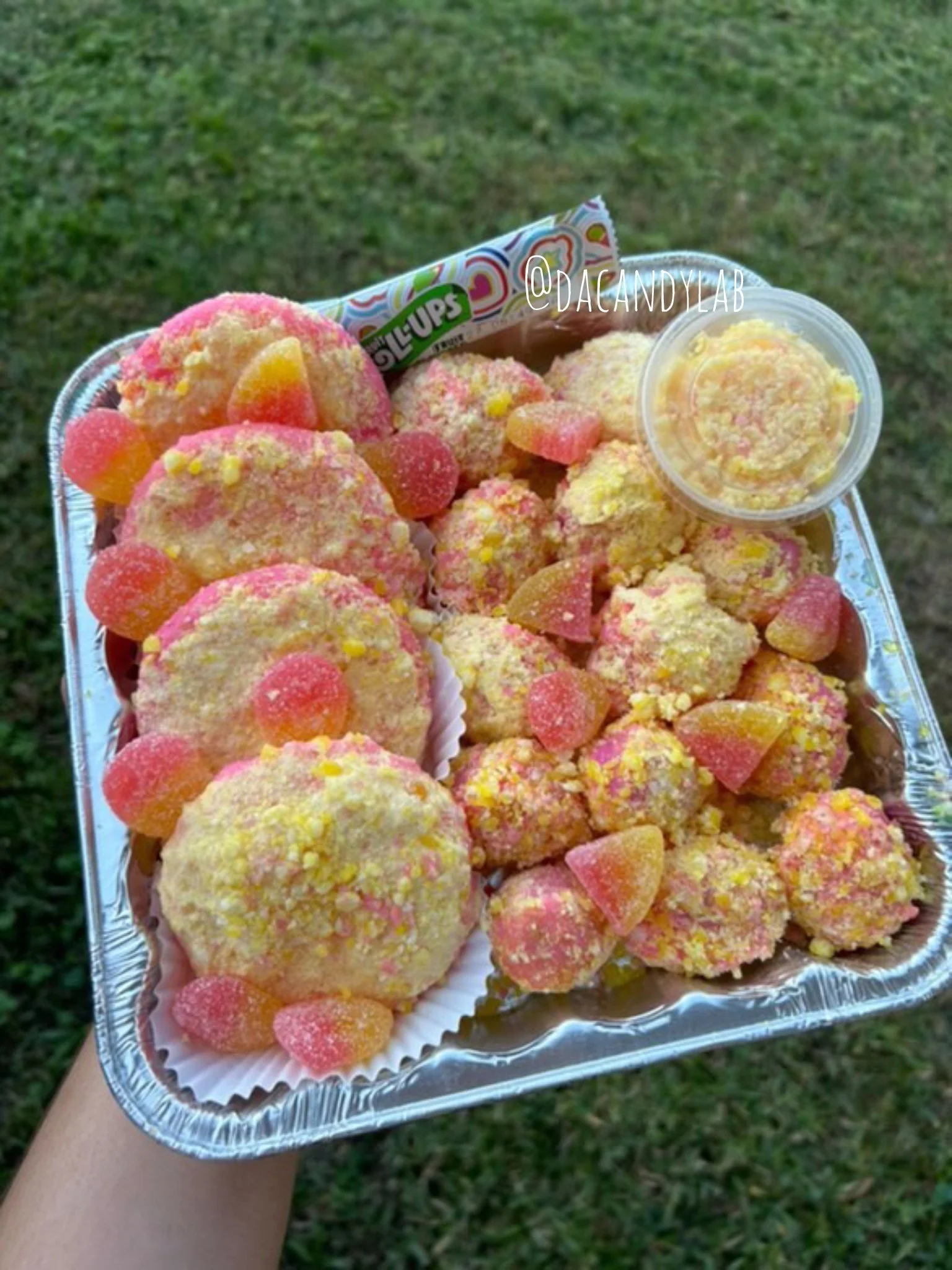 Tray of assorted colorful cookies with pink, yellow, and orange icing, a small container of sprinkles, and a pack of Skittles candies, held outdoors over grass.