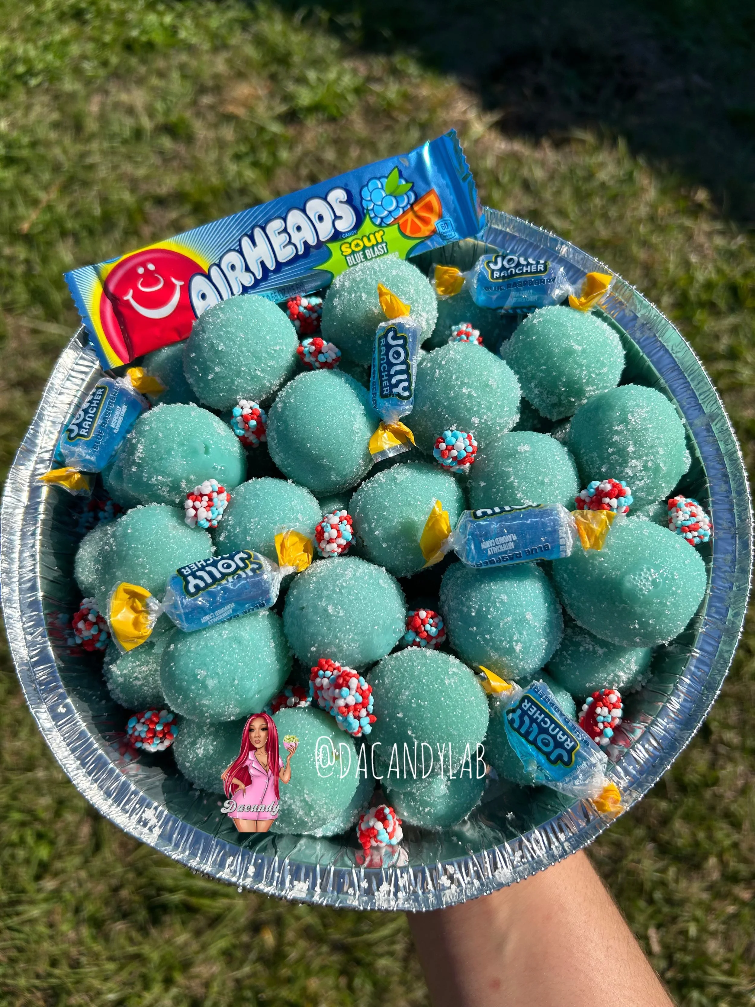 A person holding a silver tray filled with turquoise sour Airheads candies and small red and white gum balls, with a grassy background.