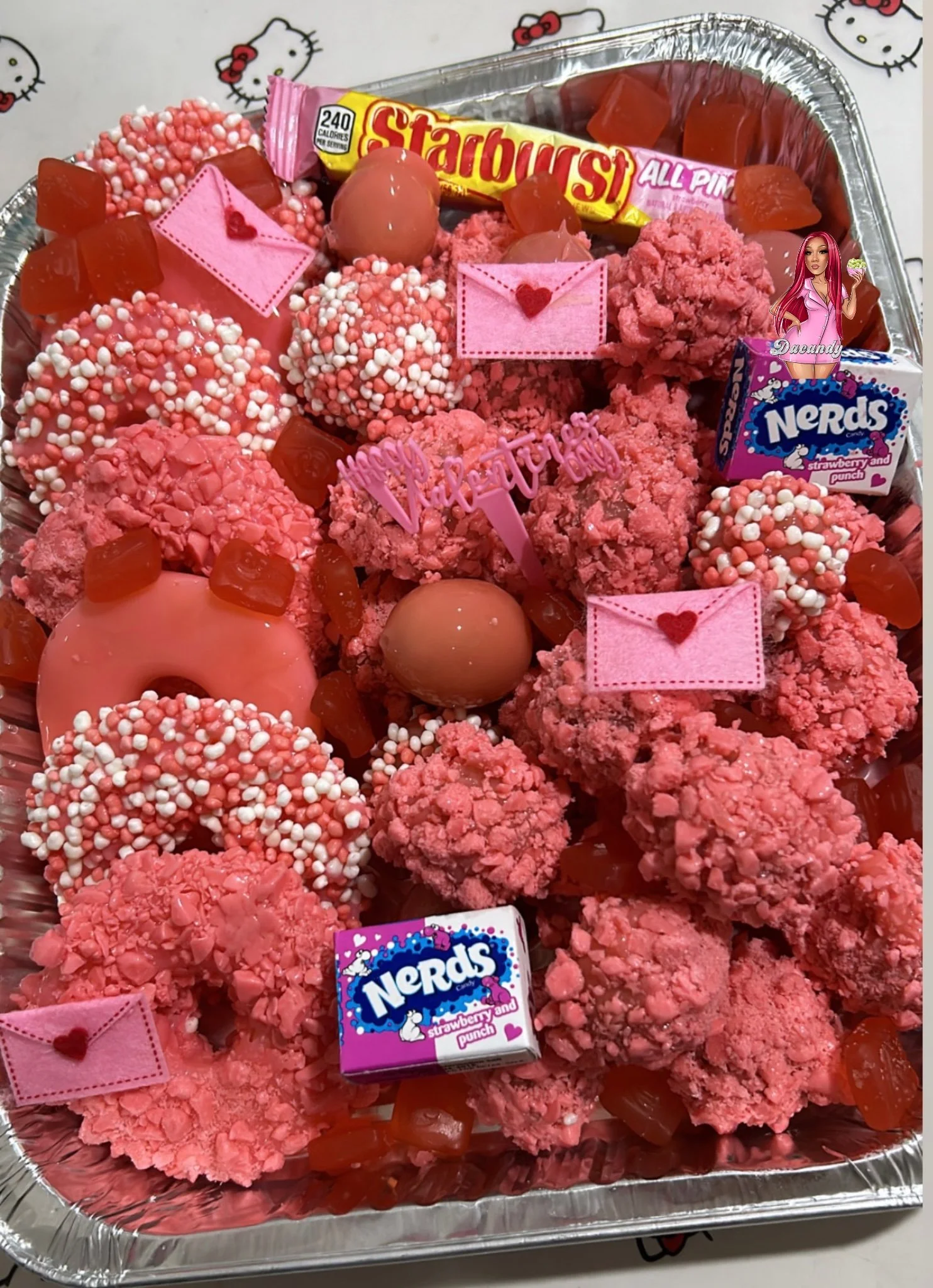 Assorted pink and red candies and treats in a foil tray, including donuts with sprinkles, chocolate-covered items, Nerds candies, and small pink decorative heart and envelope-shaped items, all on a Hello Kitty patterned tablecloth.