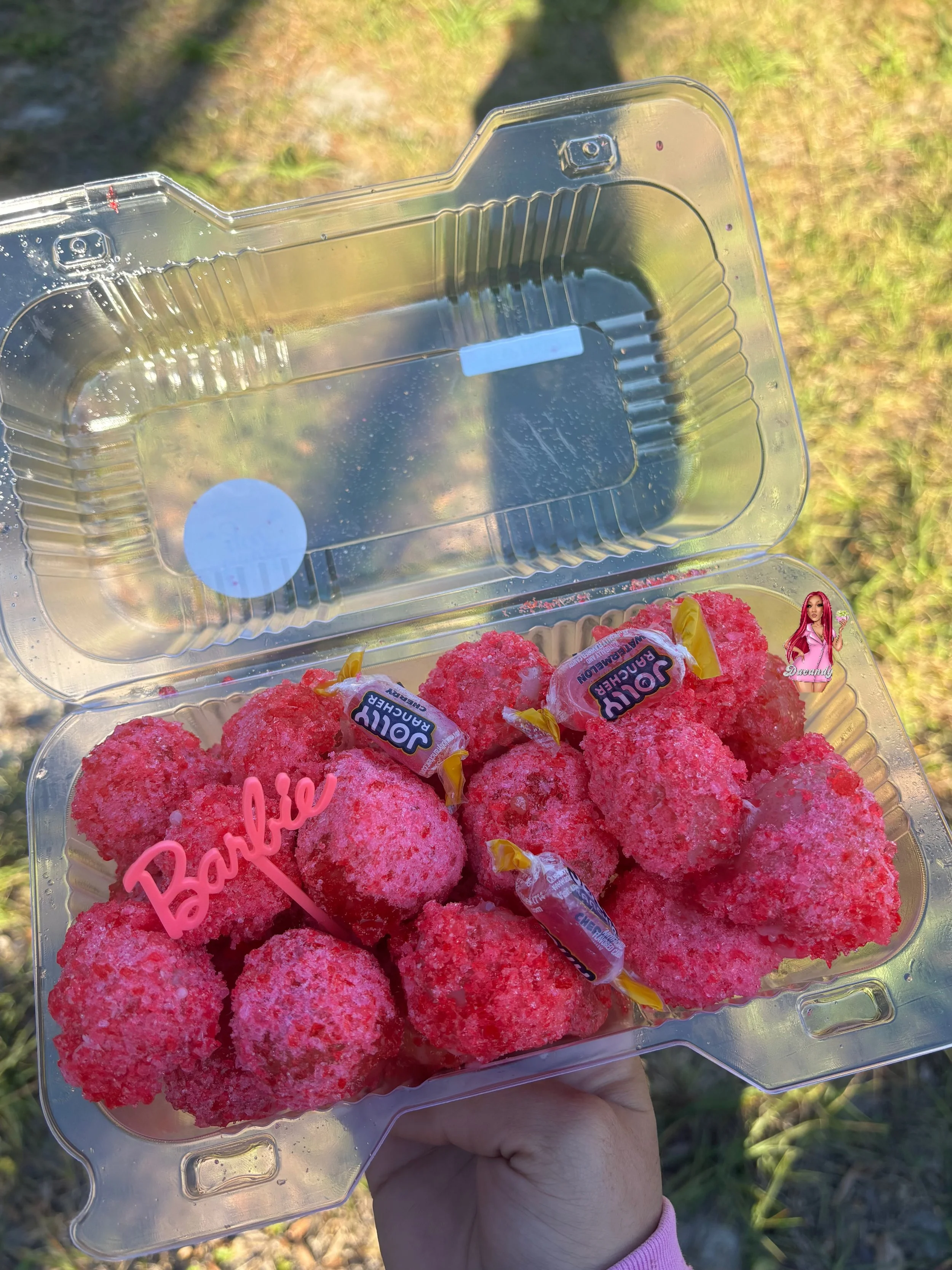 A hand holding a clear plastic container filled with pink, sugar-coated gummy candies, with some wrapped candies labeled 'JouR Rancher' inside, outdoors on grass.