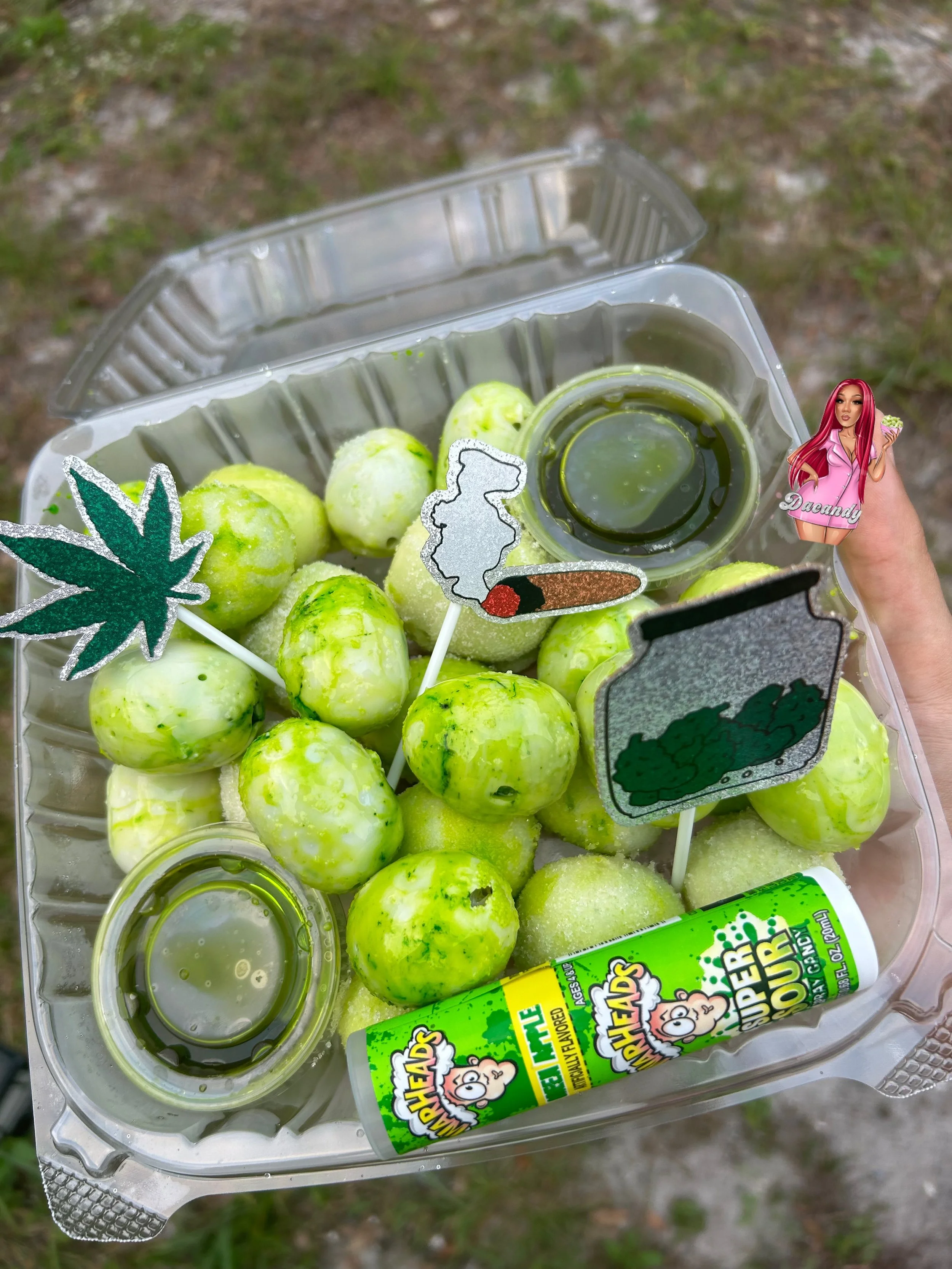 A plastic container holding green grapes, two small cups of green liquid, stickers including a marijuana leaf, a smoking joint, and a cloud near a bag of chips or snack with the label 'Dabandy,' and a tube of Flavored Lip Balm labeled 'Lemon.'
