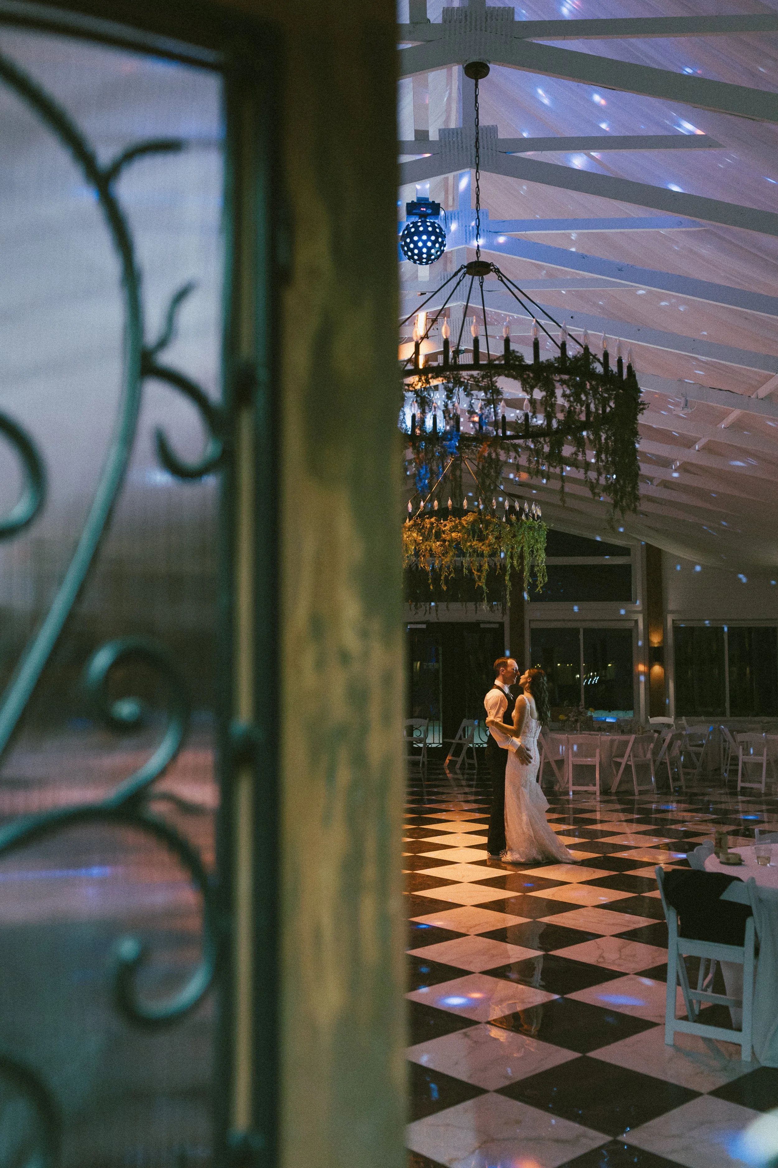 A bride and groom dancing together during their wedding reception indoors, with black and white marble floors, disco lights, floral arrangements, and the groom gazing down at the bride.