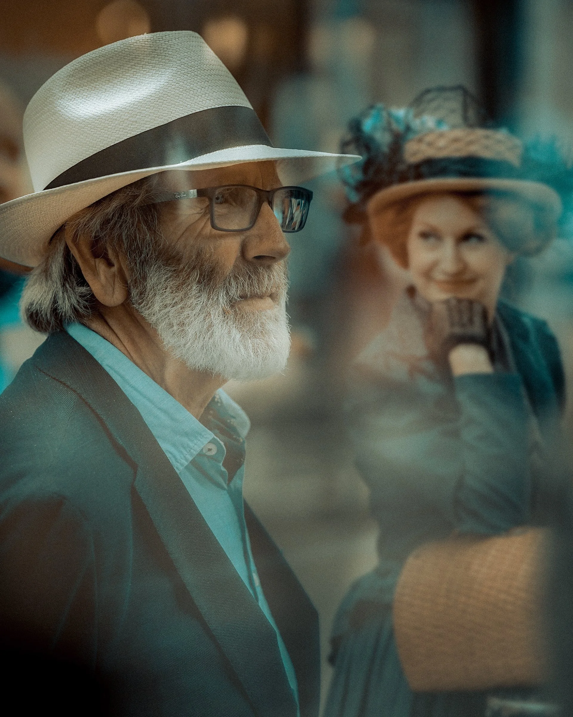 Candid Dublin street portrait of an older man in a light hat and blue jacket, with a smiling woman in a vintage hat in the background- Photo by daniya 