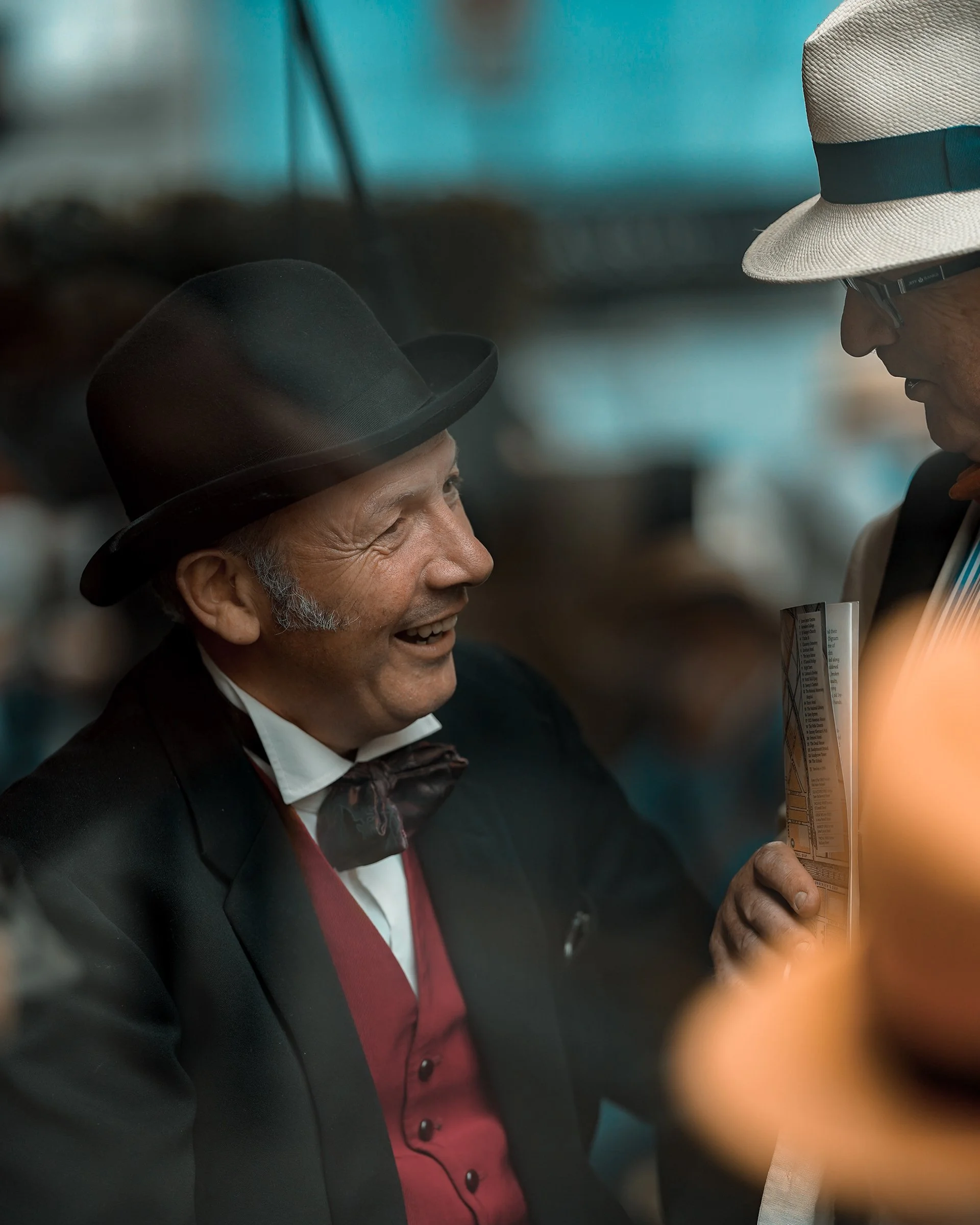Candid Dublin street portrait of a man in a black hat and bow tie, smiling while talking with another man in a white hat – Photo by Daniya, street photography.