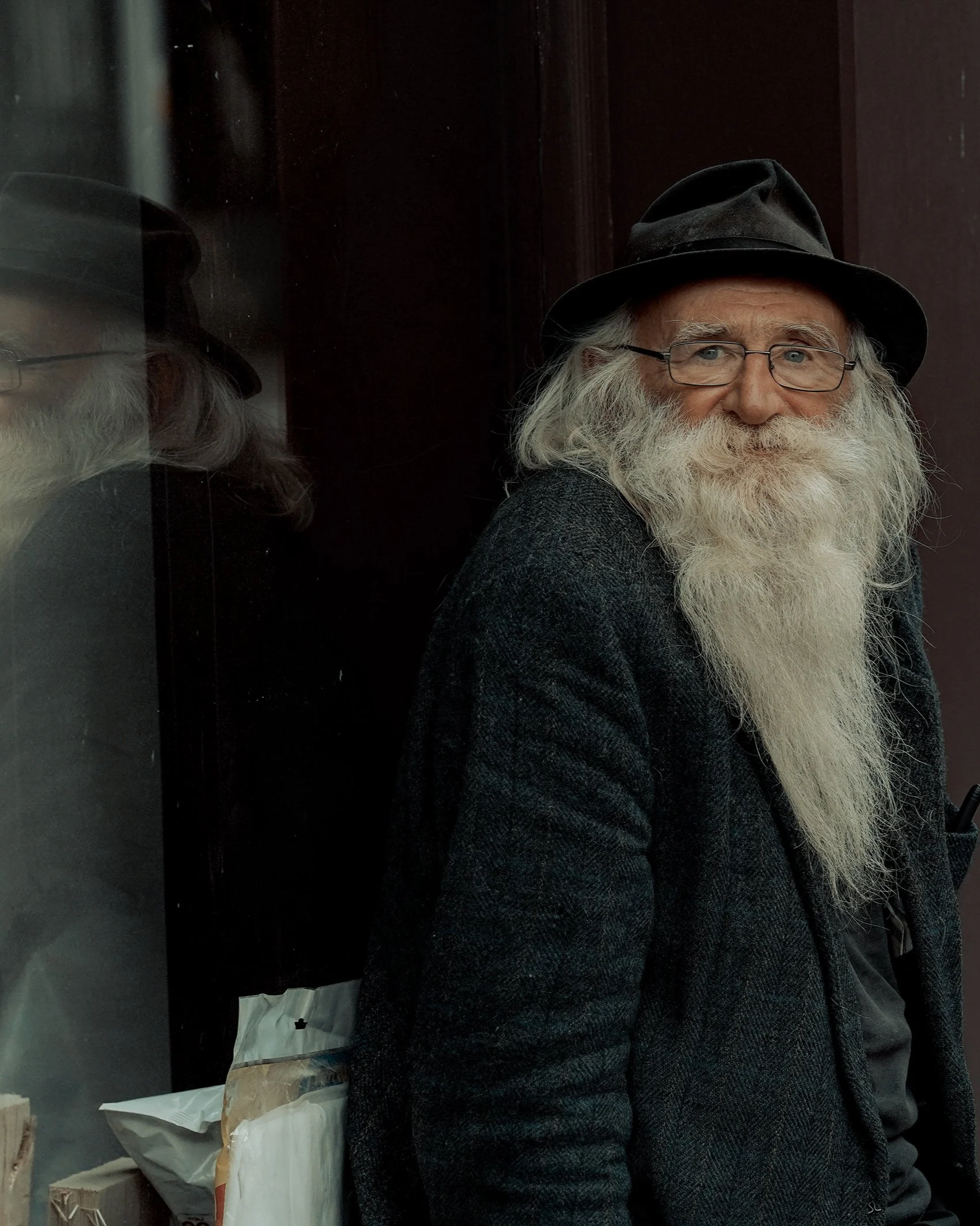 Older man with a long white beard sitting by a window, wearing a dark hat and coat, soft natural light - Dublin street photography, Photo by Daniya.