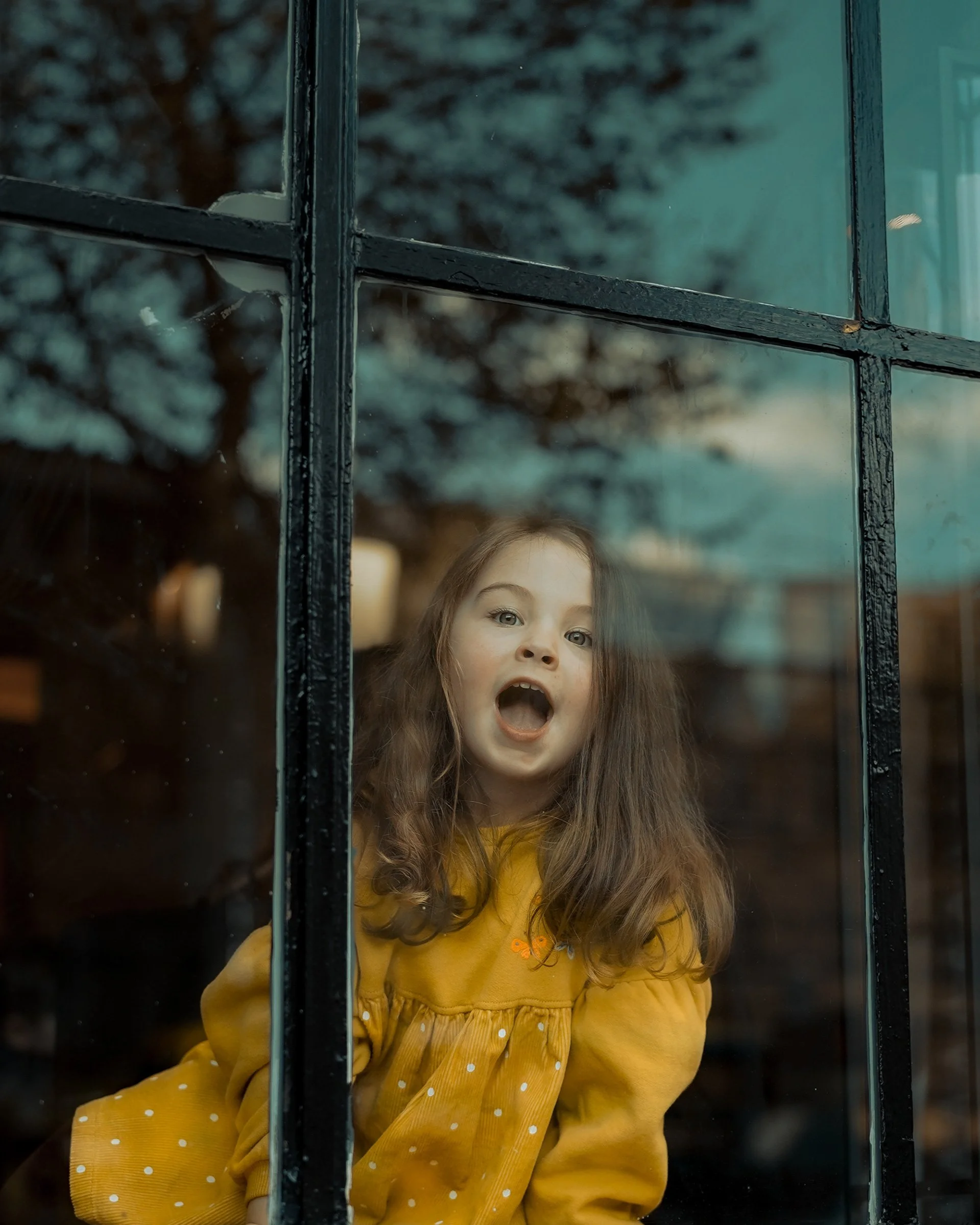 Little girl in a yellow coat looking out through a window, playful and candid — Dublin street photography, Photo by Daniya.