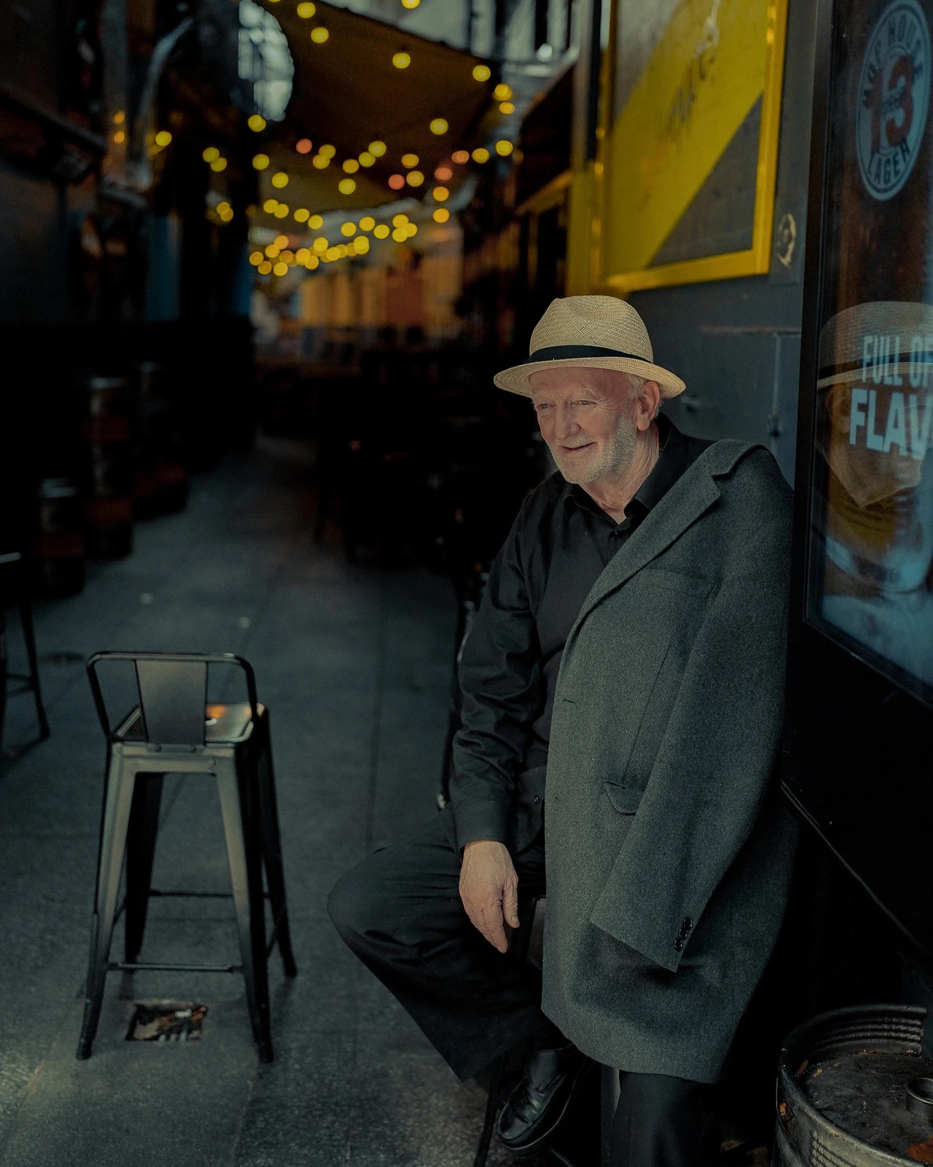 Older man smiling in an alleyway with string lights, wearing a beige hat and dark coat — Dublin street photography, Photo by Daniya.