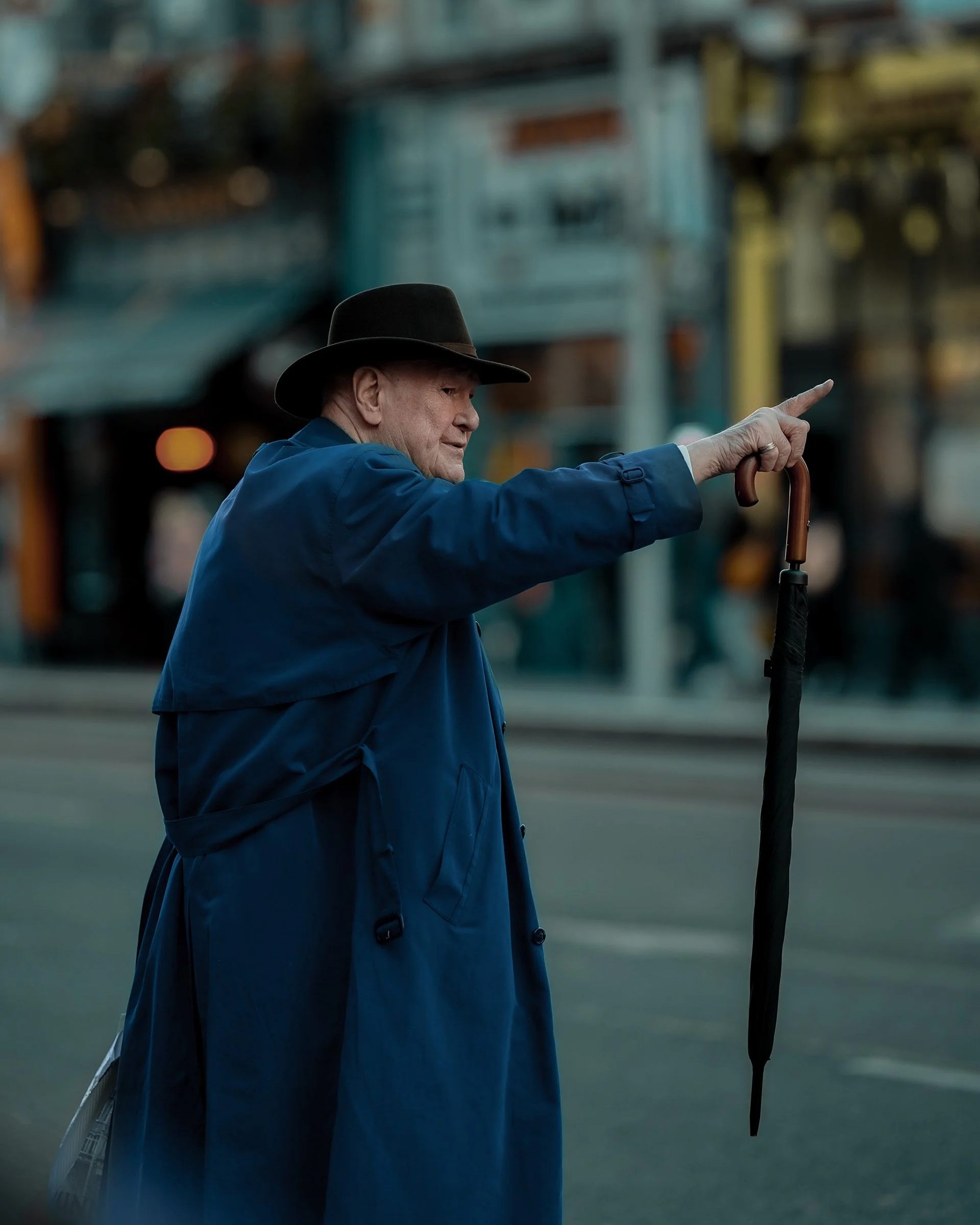 Older man in a blue coat and black hat holding an umbrella and gesturing across the street — Dublin street photography, Photo by Daniya.