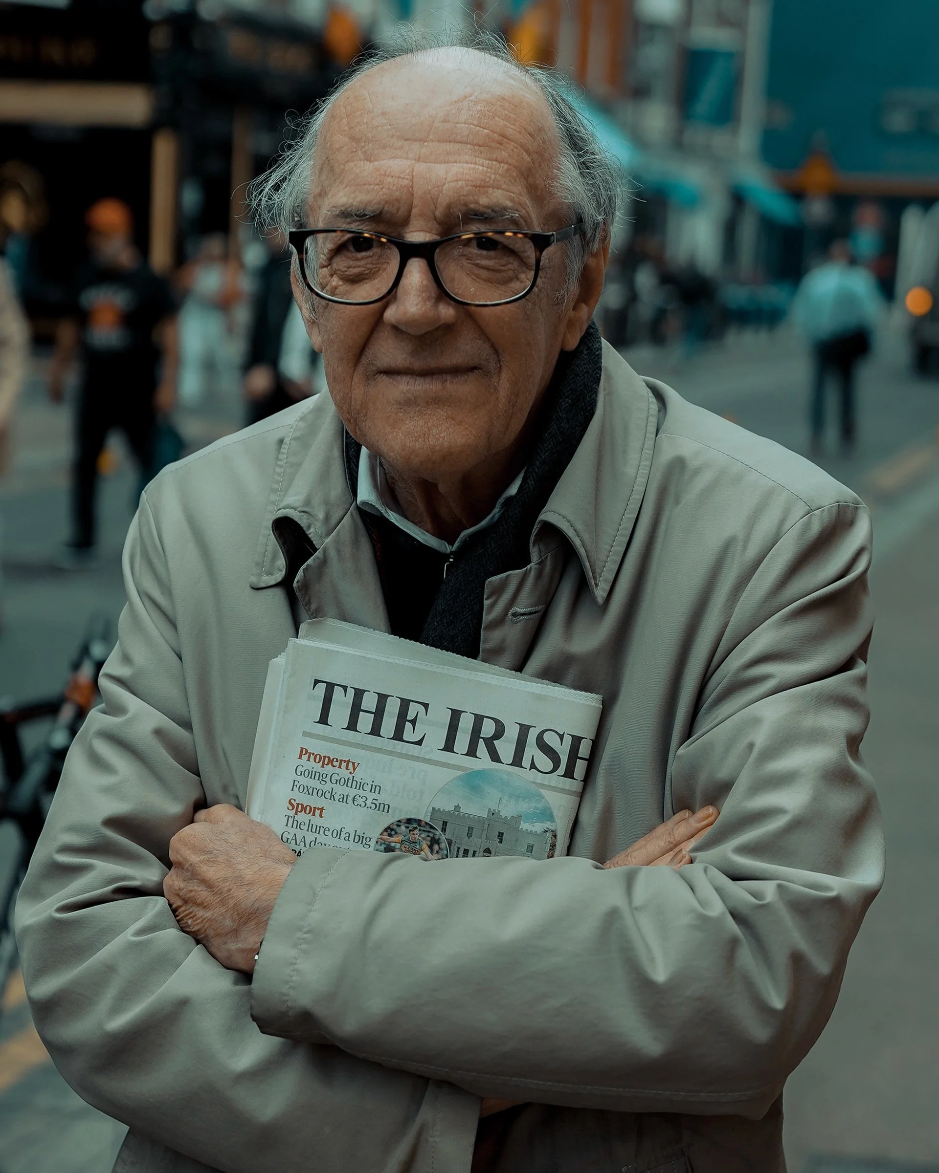 Older man in a beige coat holding a folded Irish newspaper, candid urban portrait — Dublin street photography, Photo by Daniya.
