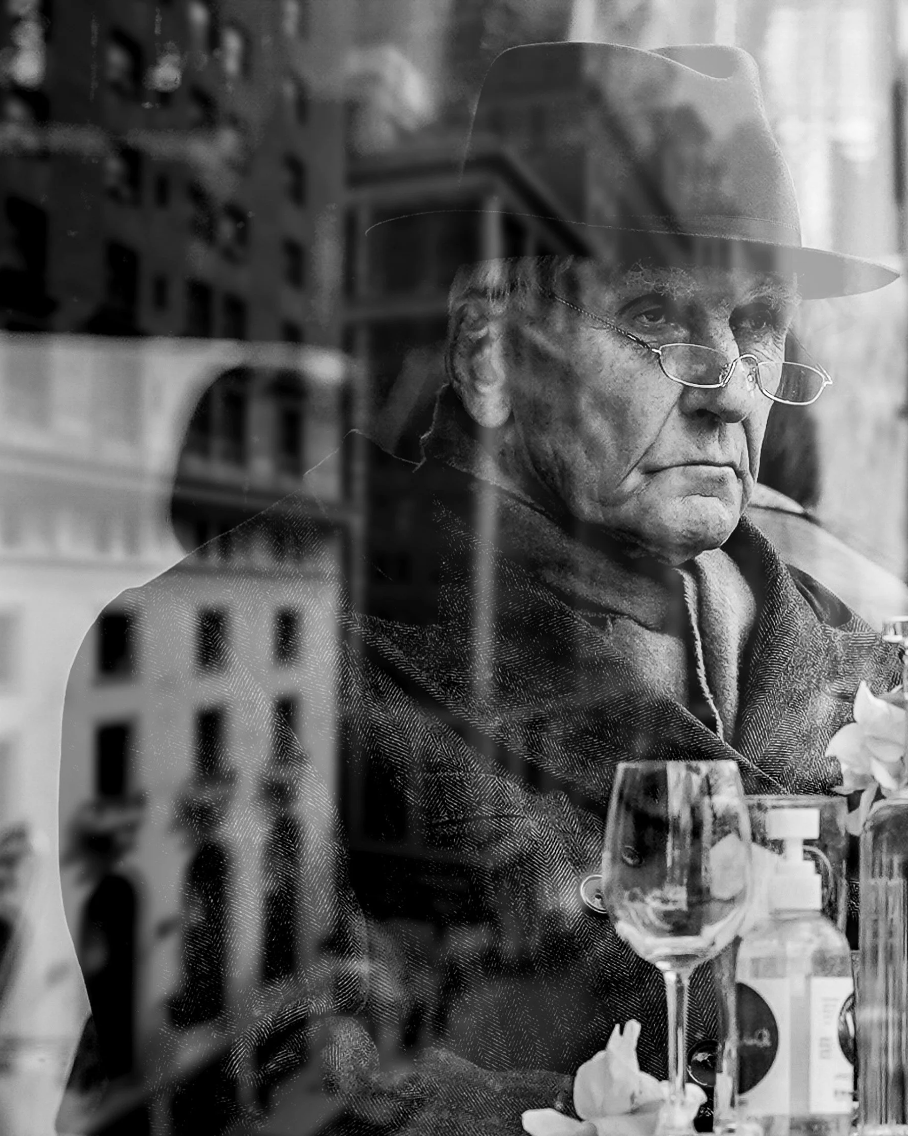 Moody black-and-white candid of an older man in a café with wine glasses, seen through the window reflection — Dublin street photography, Photo by Daniya.