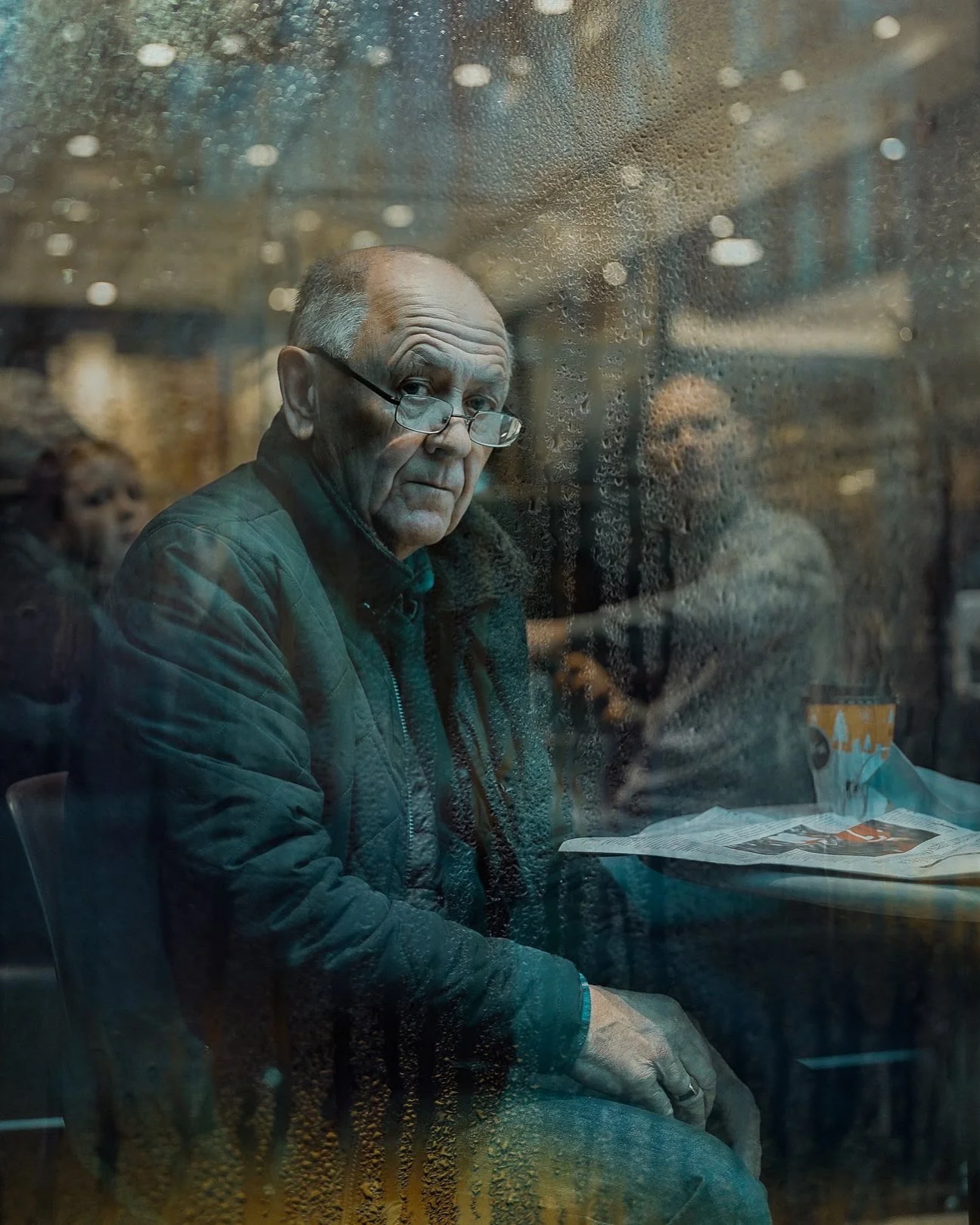 Older man in glasses sitting by a café window on a rainy day, moody reflection — Dublin street photography, Photo by Daniya.