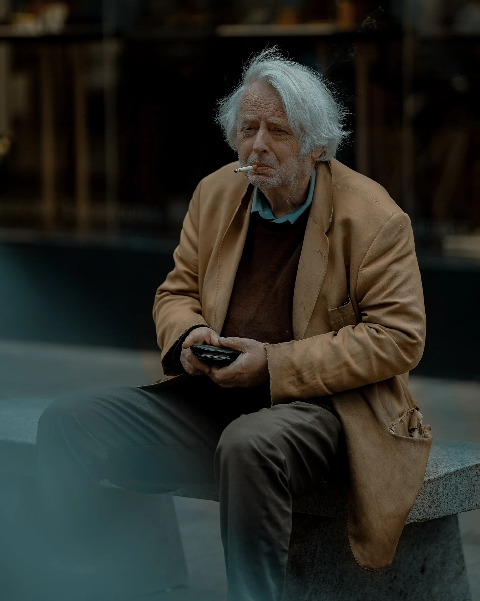 Street portrait of an elderly man with white hair, sitting on a bench in Dublin, wearing a mustard coat and smoking, documentary street photography.