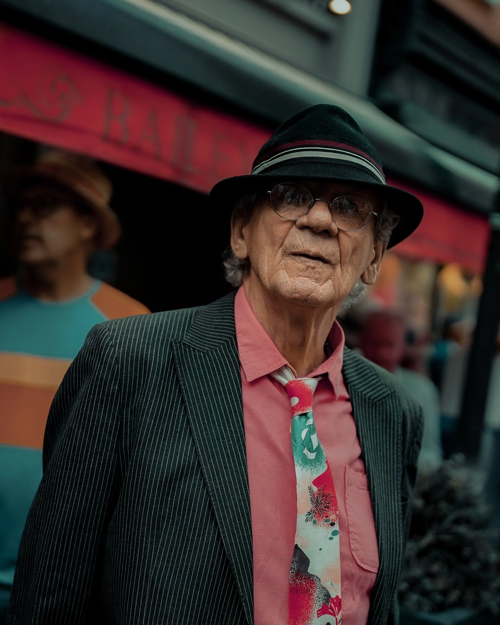 Street style portrait of an older man in a striped suit jacket, pink shirt, and tie, wearing a black hat, captured in Dublin, street photography.