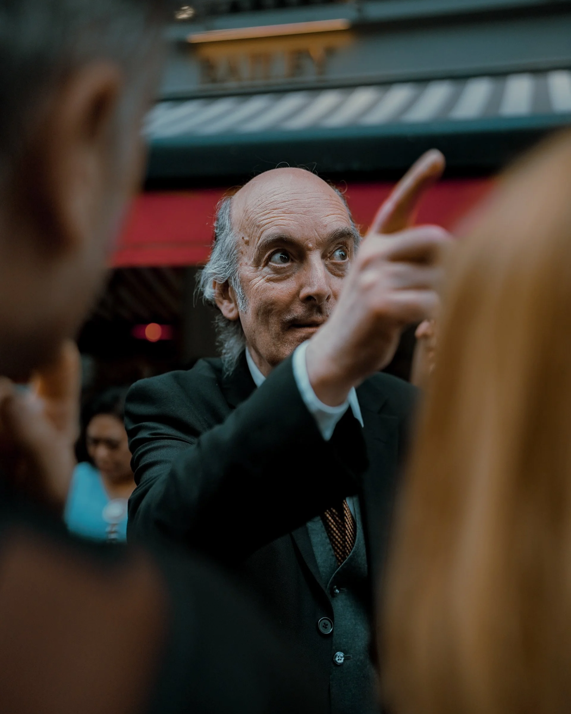 Candid street portrait of a bald elderly man in a suit gesturing with his finger, surrounded by a crowd in Dublin.