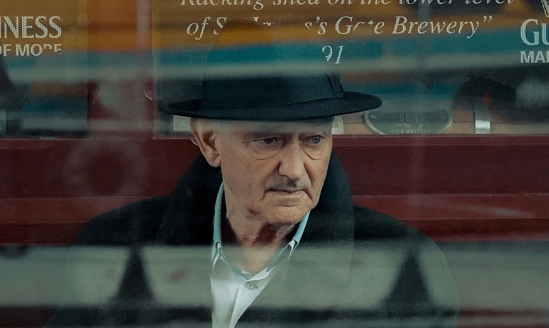 Older man in a dark hat and coat gazing out from a pub window with a Guinness sign behind — Dublin street photography, Photo by Daniya.