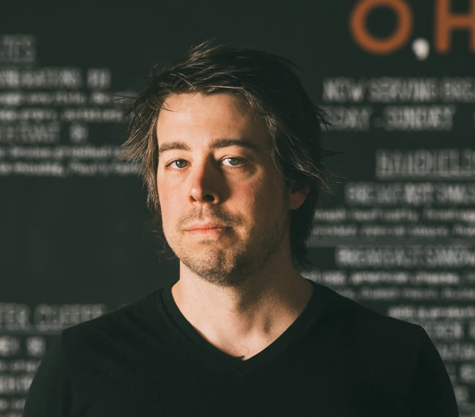Portrait of a young man with medium-length dark hair, light skin, and a slight beard, wearing a black shirt, standing in front of a blurred blackboard with white text.