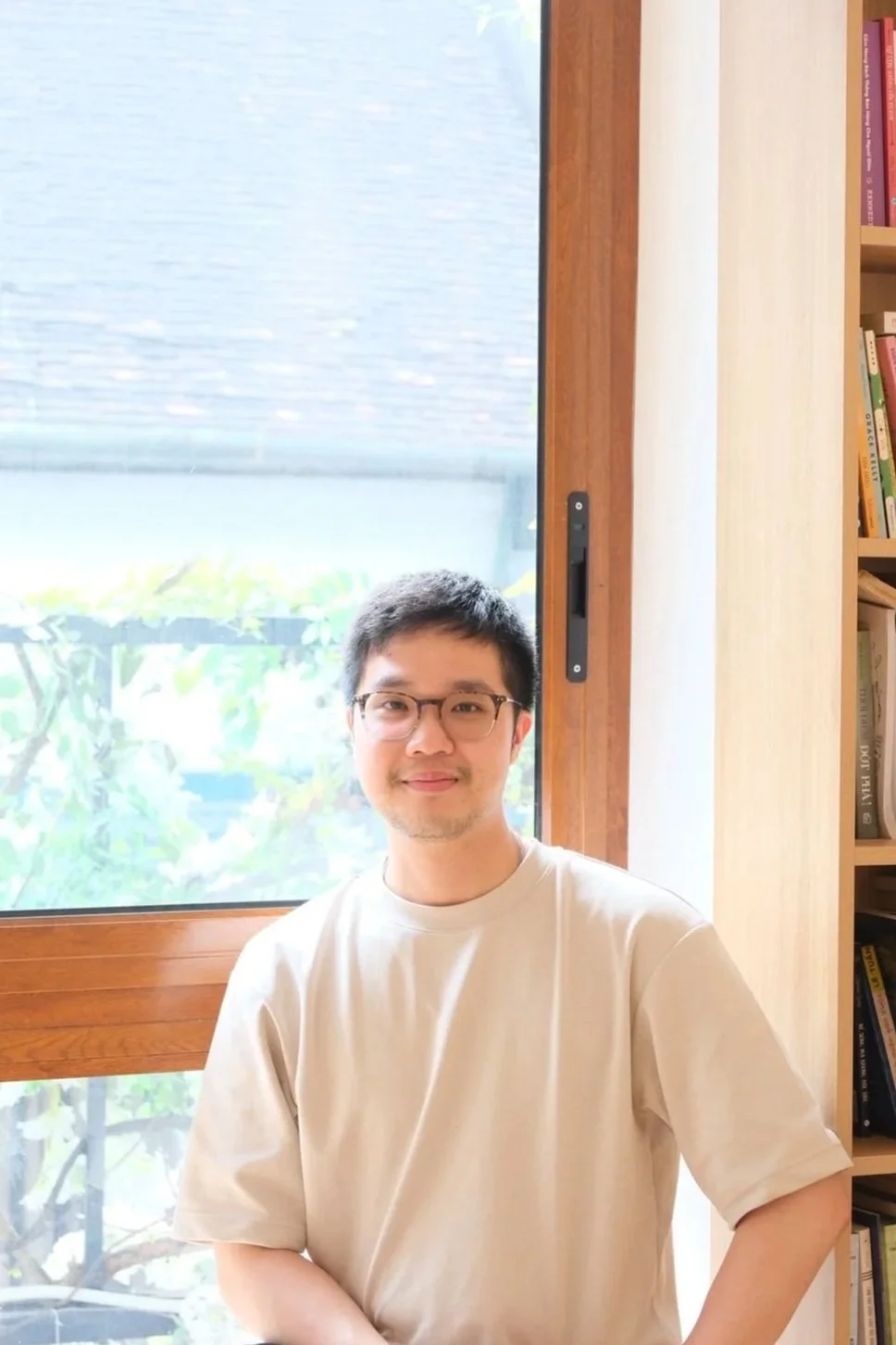 A man with glasses and a beige t-shirt standing indoors near a window with greenery outside, a bookshelf to his right.