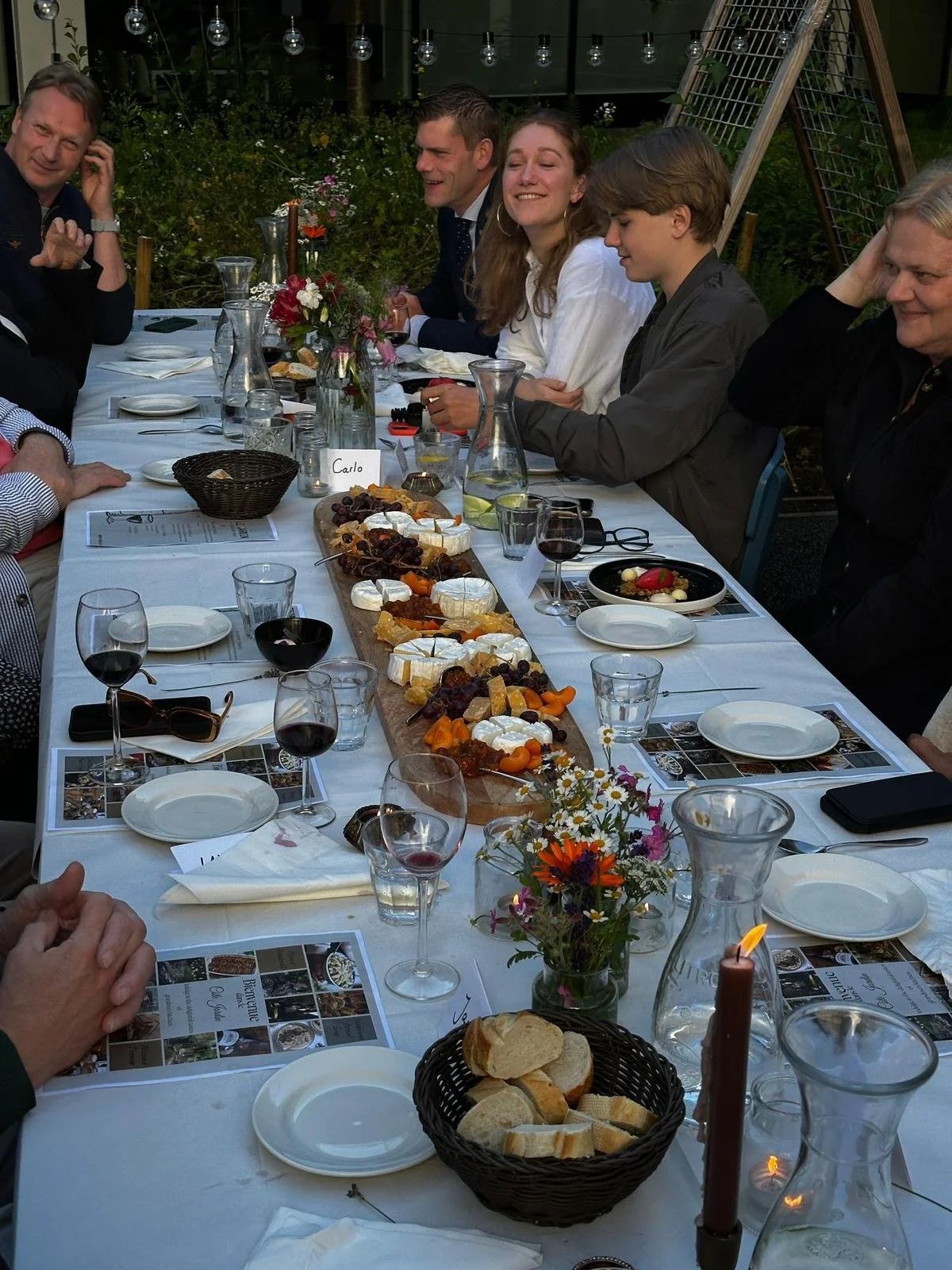 People sitting at a long outdoor dining table enjoying a meal with a cheese platter, glasses of wine, and floral decorations.