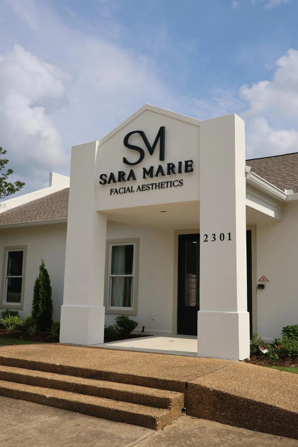 Exterior view of a facial aesthetics clinic named Sara Marie, with a white building, black signage, and stairs leading to the entrance, under a partly cloudy sky.