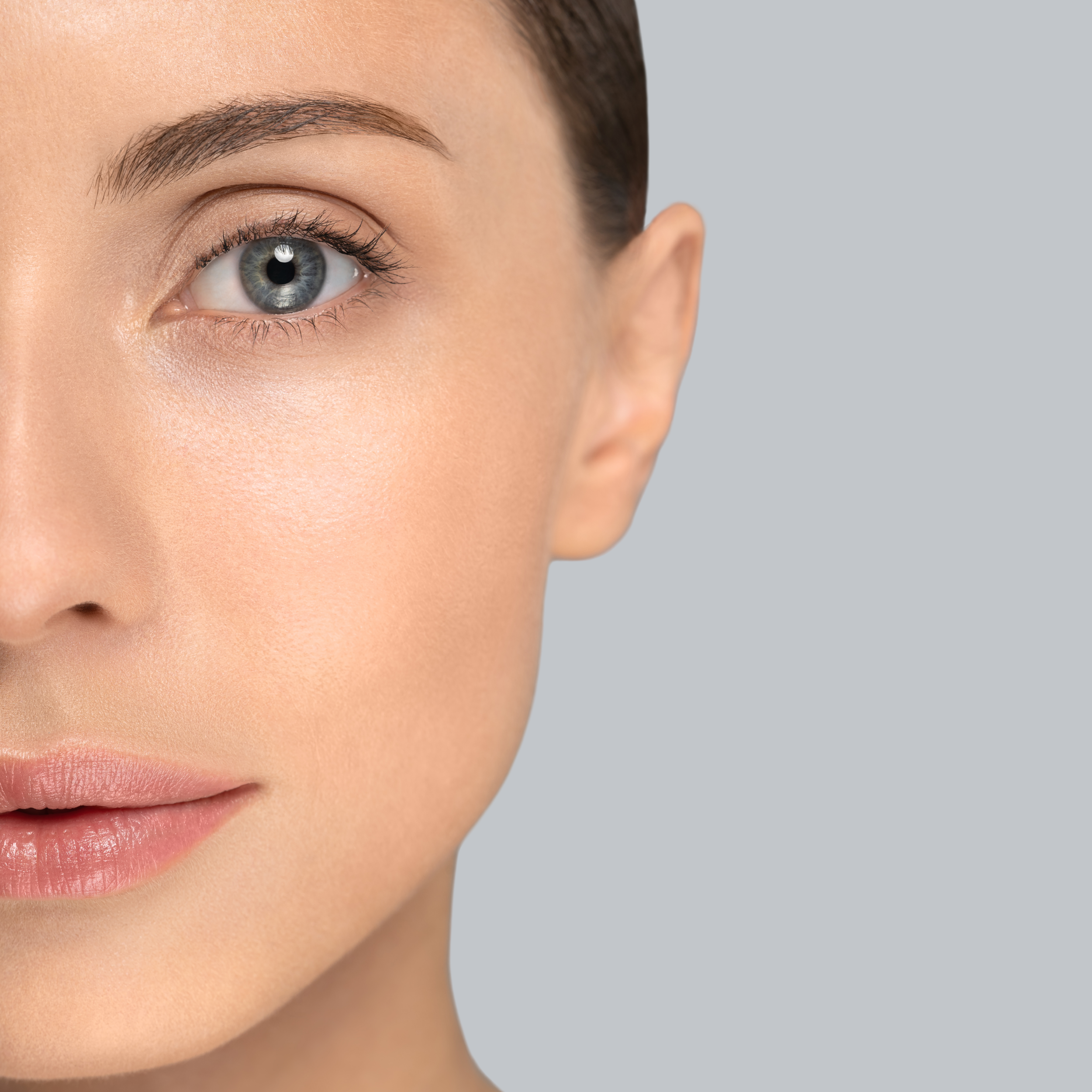 Nahaufnahme eines Frauengesichts mit blauen Augen, geglätteter Haut und natürlichem Make-up, vor hellem grauem Hintergrund.