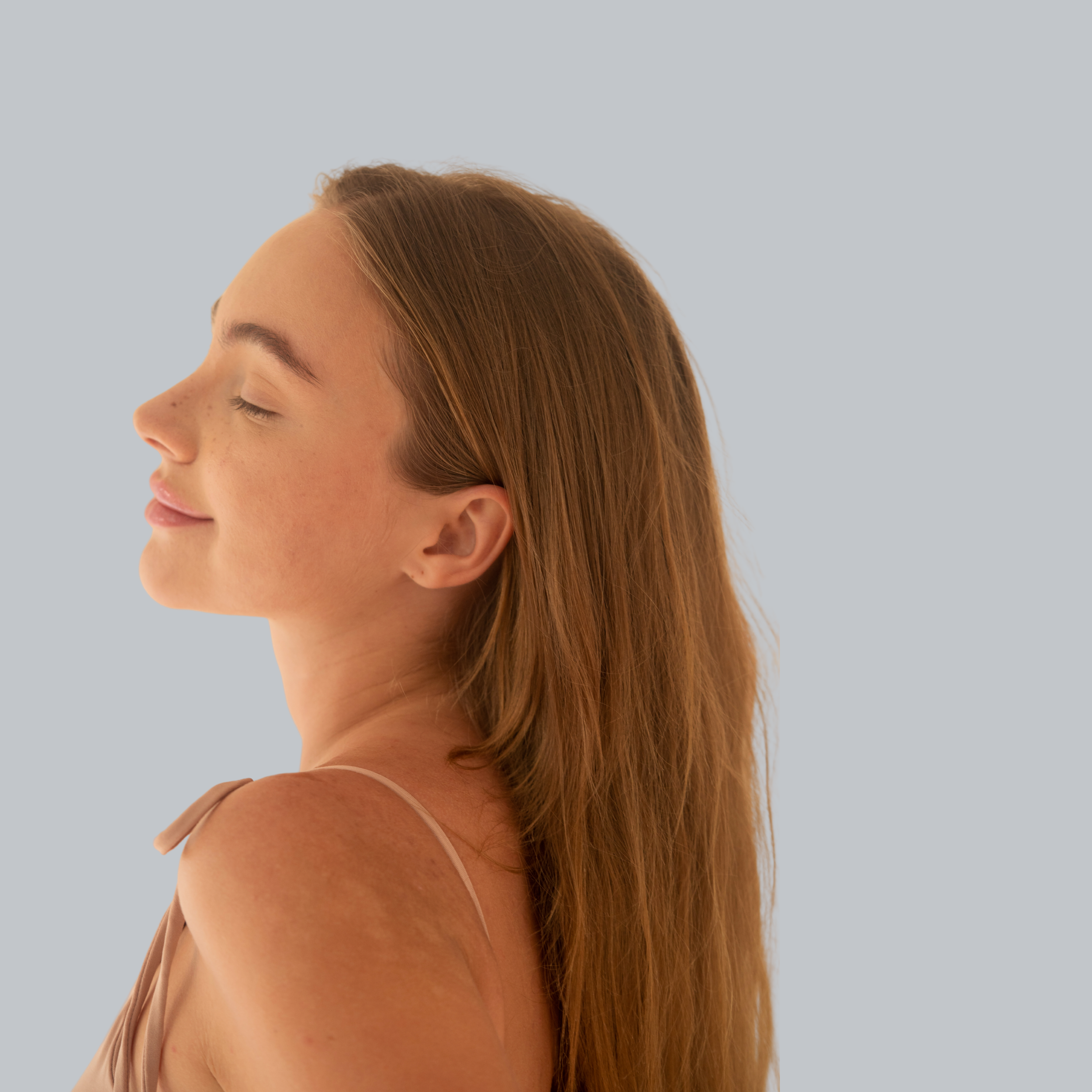 Nahaufnahme einer lächelnden roten-haired Frau mit geschlossenen Augen, seitlicher Blick, lächelt entspannt, hat seidige, lange Haare, trägt einen nudefarbenen Top, Hintergrund in Hellgrau.
