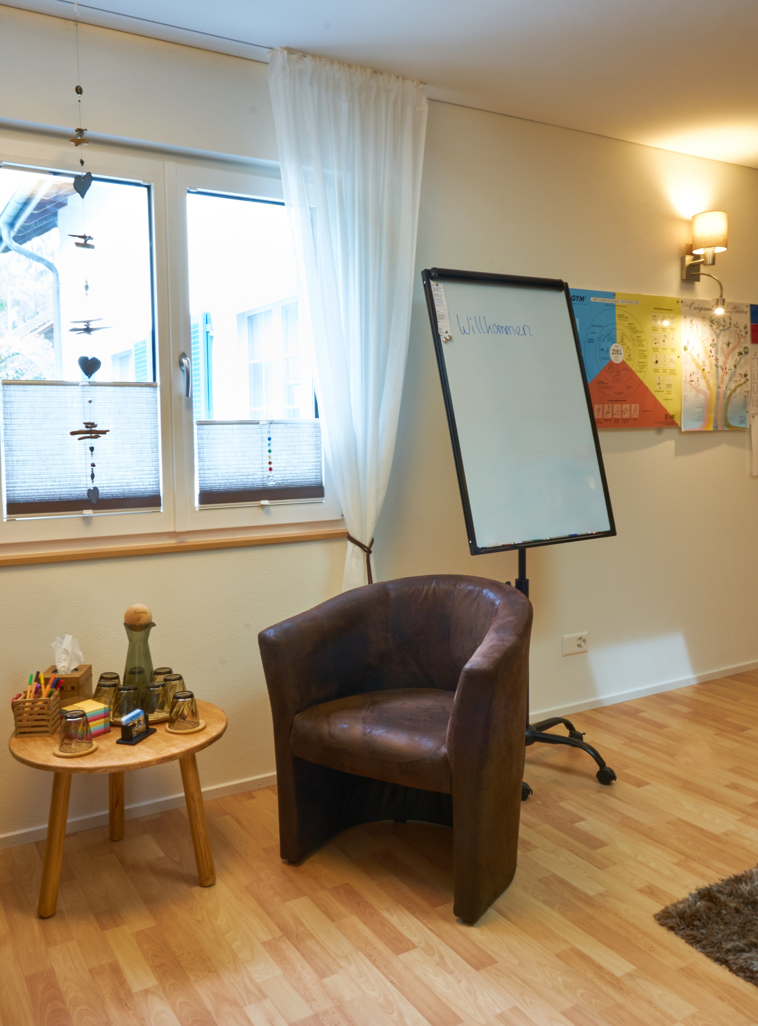 Ein gemütlicher Raum mit einem Fenster, einer braunen Ledersessel, einem kleinen runden Holztisch mit bunten Stiften und Gläsern, einem weißen Stehtafel mit der Aufschrift 'Willkommen' und einer Wanddekoration mit einem Diagramm.