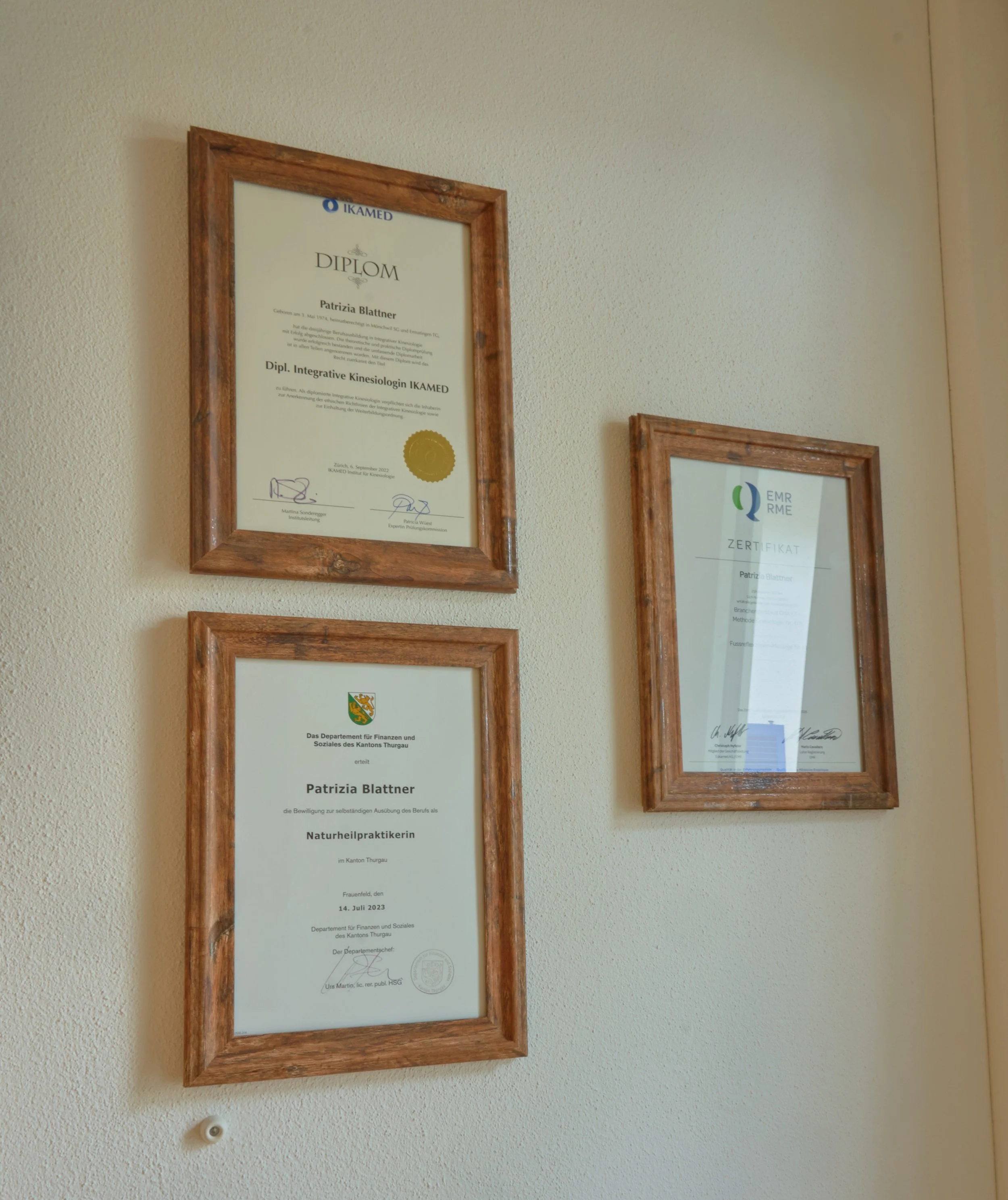 Drei Diplome und Zertifikate an einer Wand in Holzwandrahmen, die Qualifikationen der Person in Kinesiologie, Naturheilkunde und Sozialwissenschaften anzeigen.