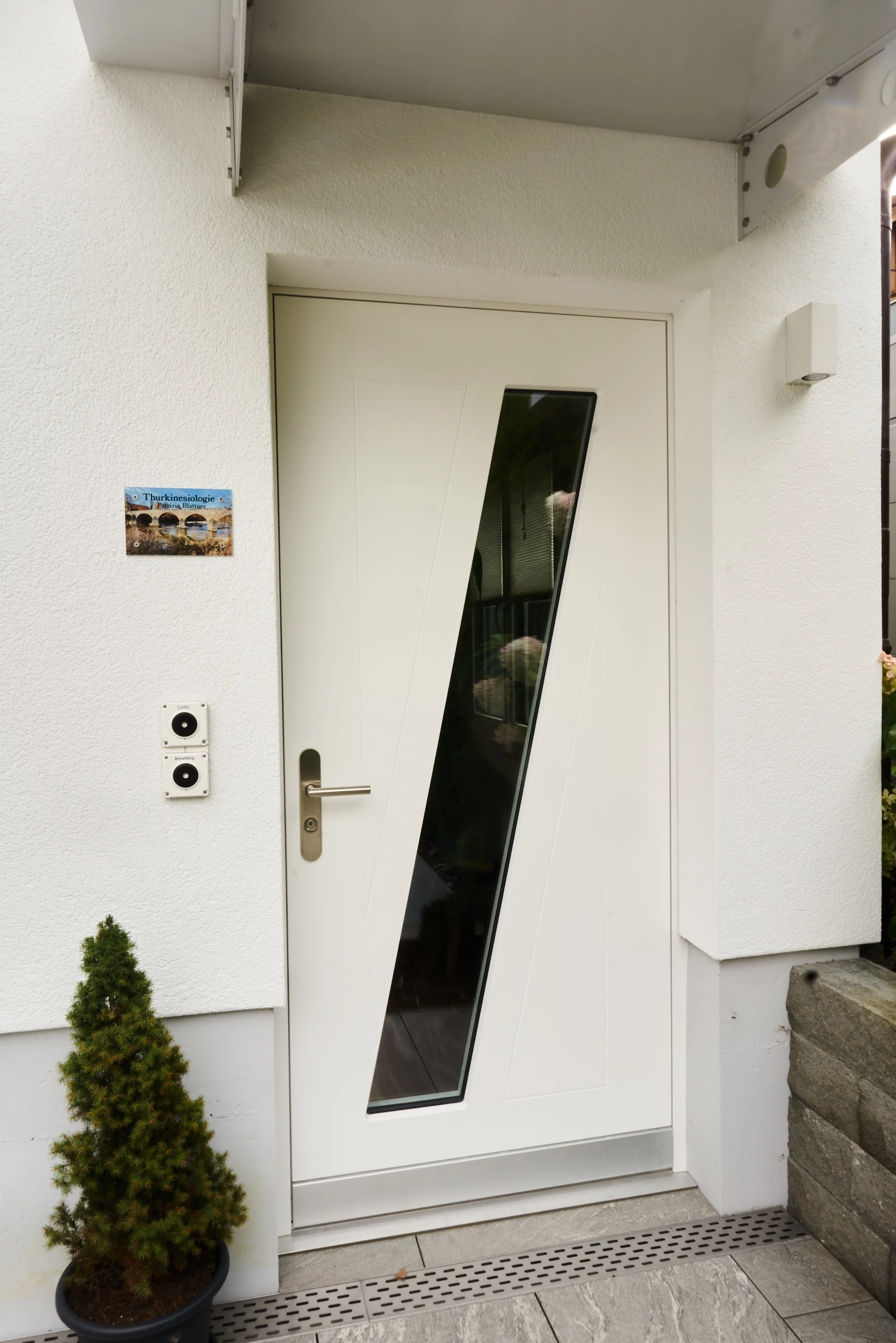Moderner Eingang mit weißen Tür, diagonalem Fenster, einer kleinen Pflanze und einer Hausnummer