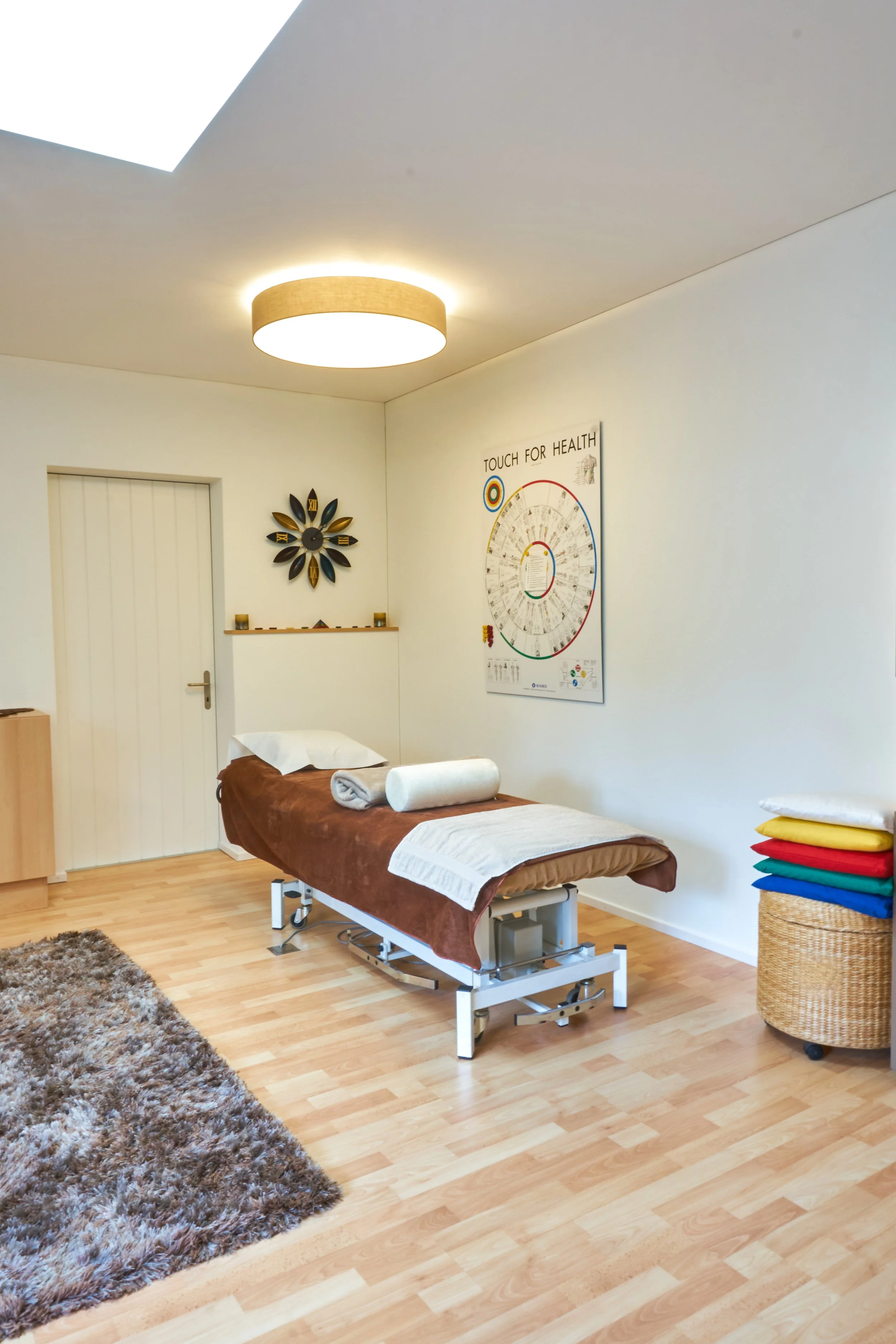 Behandlungsraum mit Behandlungsliege, bunten Decken auf einem Wäschekorb, Wanddekoration und Poster an der Wand, Holzfußboden, Teppich, Lampen an der Decke.