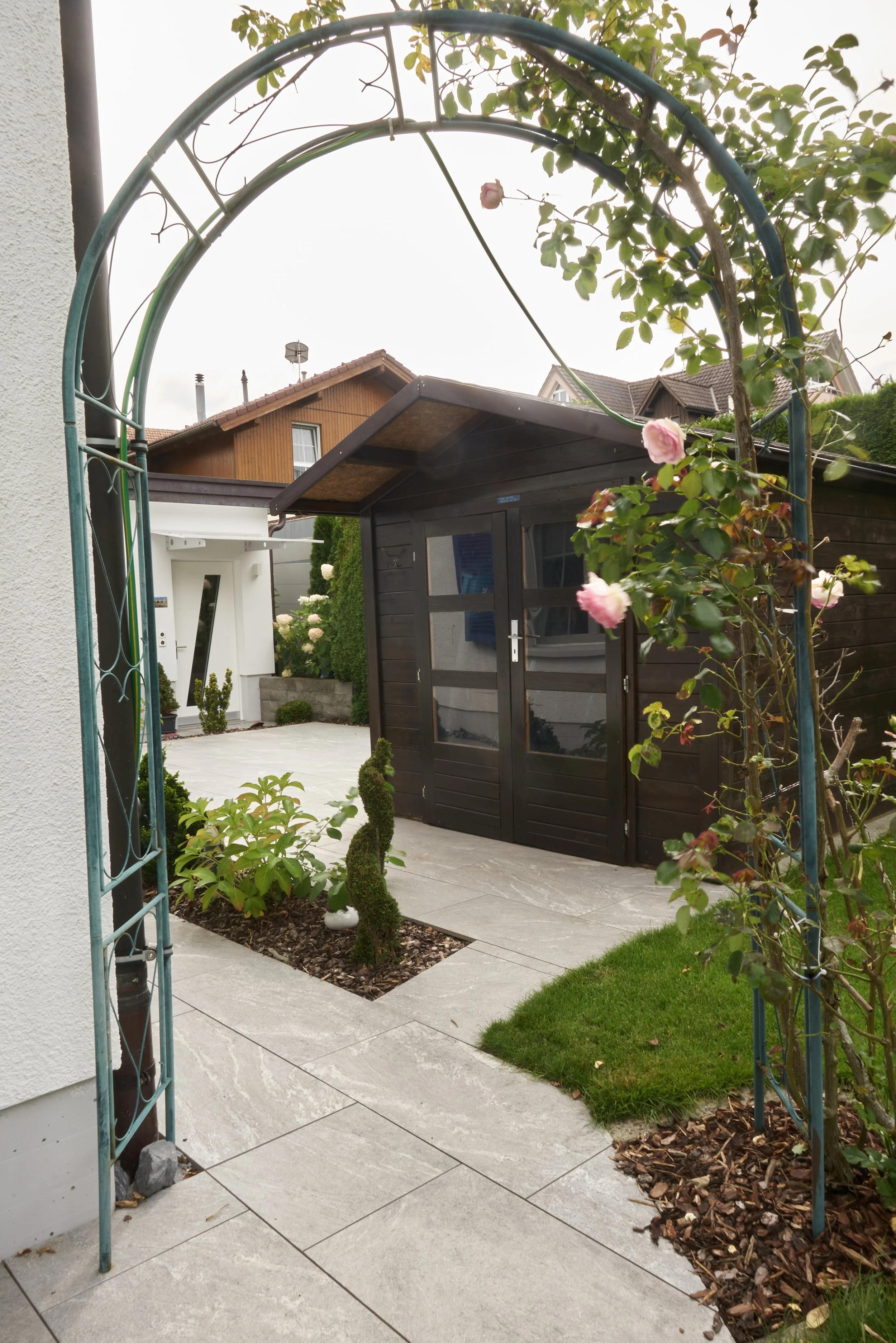 Garten mit Topfpflanzen, einem kleinen Gartenhäuschen und einem modernen Haus im Hintergrund.