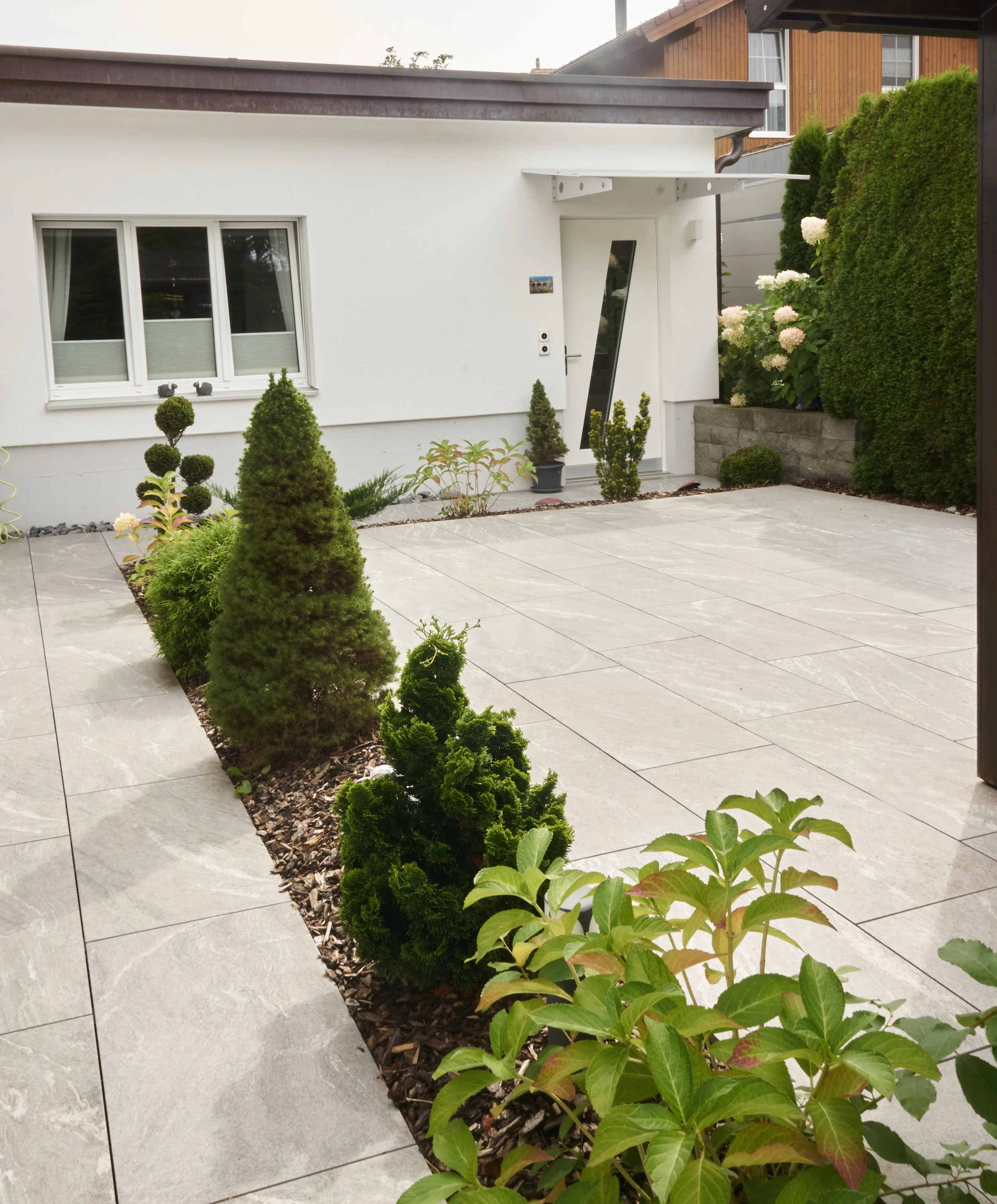 Ein gepflegter Garten vor einem weißen Haus mit modernen Fenster und Tür. Im Vordergrund sind mehrere kleine, grüne, kunstvoll geschnittene Sträucher entlang eines Kiesstreifens. Im Hintergrund sind große, grüne Sträucher und blühende Hortensien zu sehen.