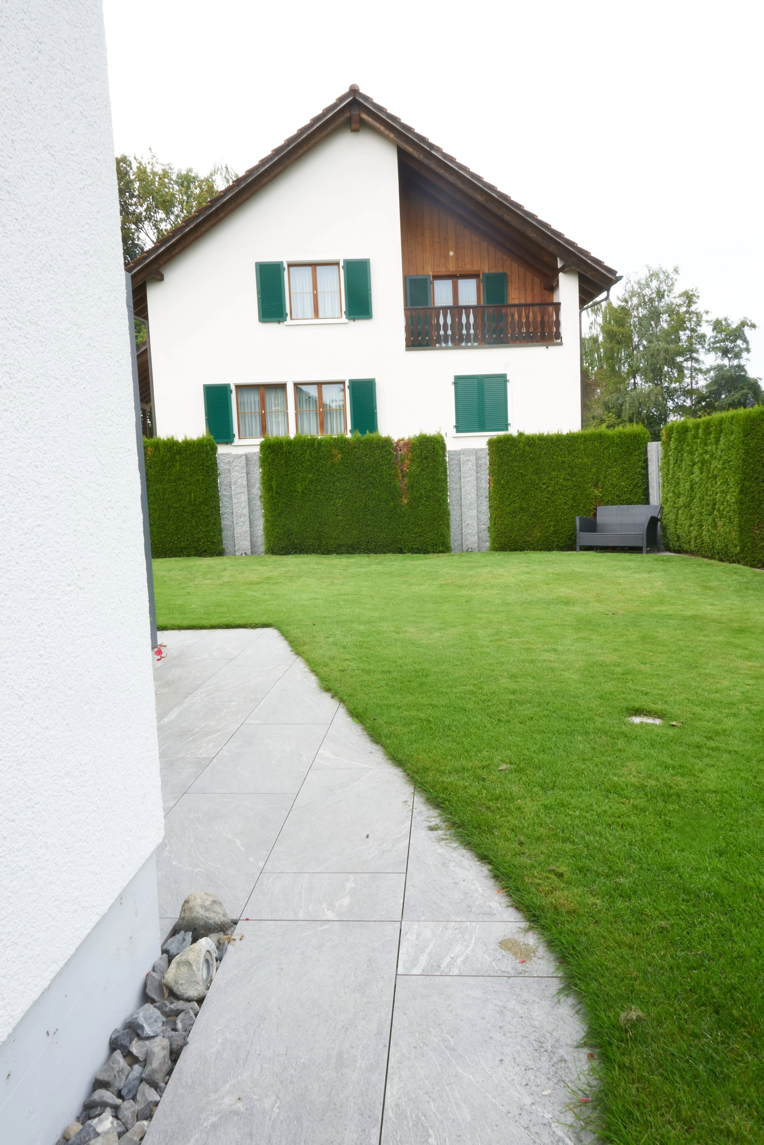 Garten mit grünem Rasen, Hecken und einem schwarzen Sitzmöbel vor einem weißen Haus mit grünen Fensterläden und einem Balkon.