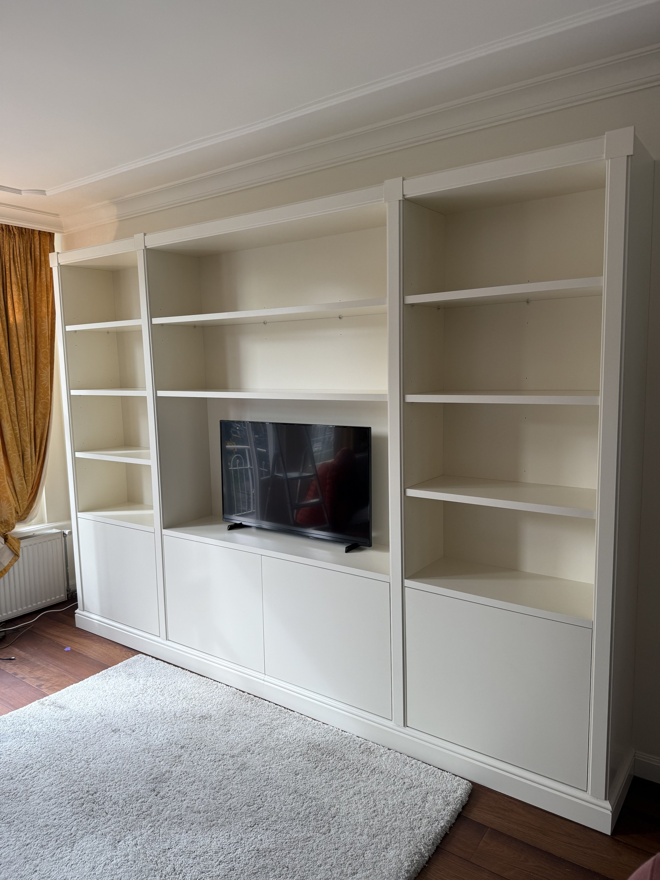 Witte boekenkast met een televisie in het midden, een paar planken en laden, in een woonkamer met houten vloer, witte muur en oranje gordijnen.