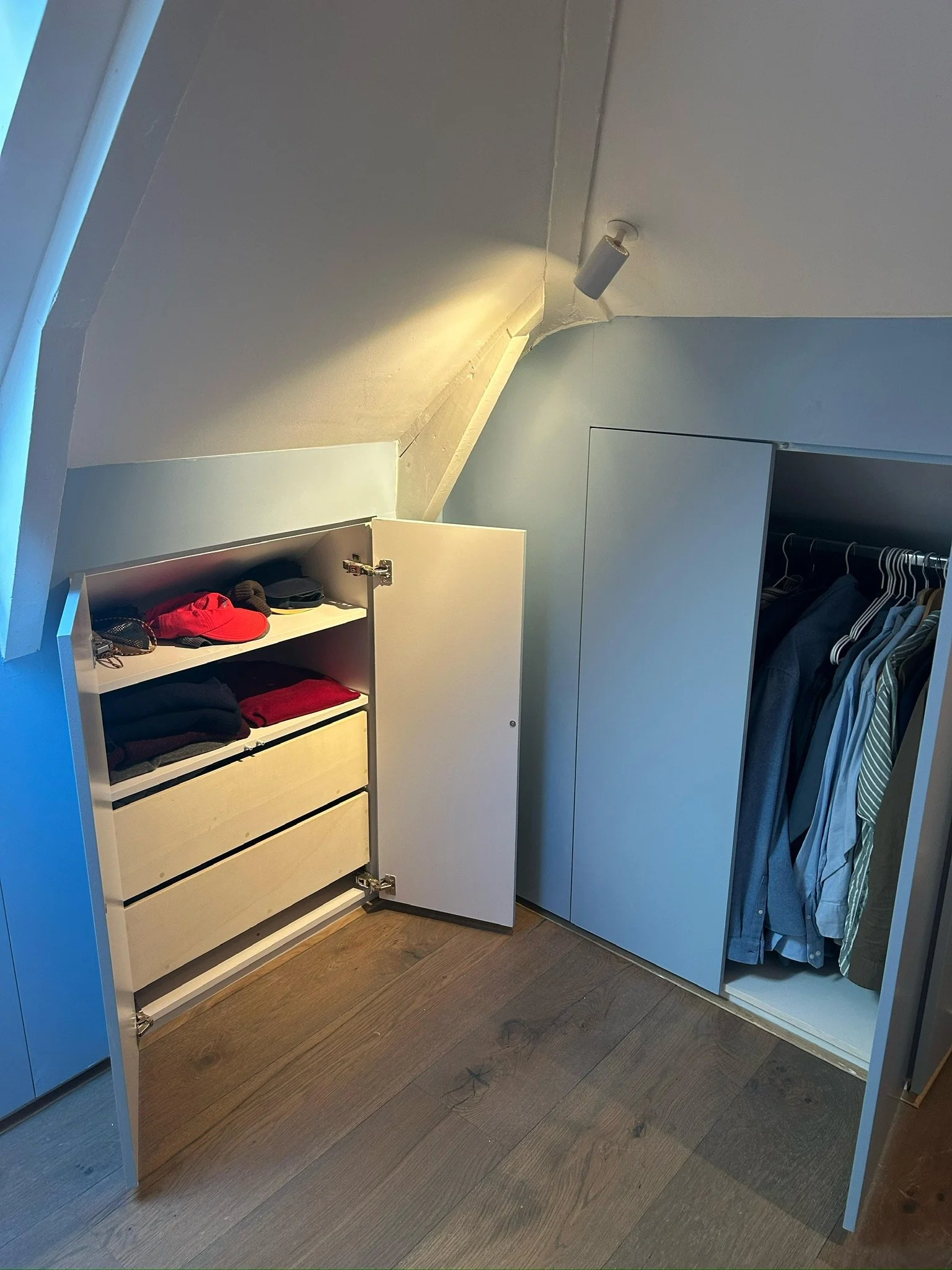 Ingericht klein kastje met kleding en accessoires, met open deurtjes, onder een schuine plafondhoek, naast een kledingkast met hangende kleding.