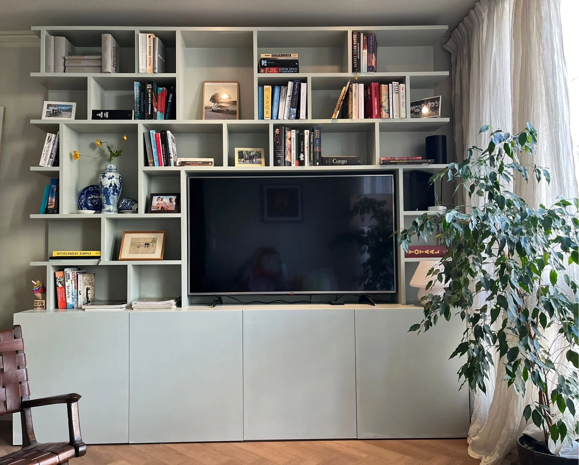 Een witte muur met een grote televisiescherm, omgeven door een witte boekenplank met boeken, foto's en decoraties, en een grote groene plant aan de rechterkant, naast een raam met witte gordijnen.