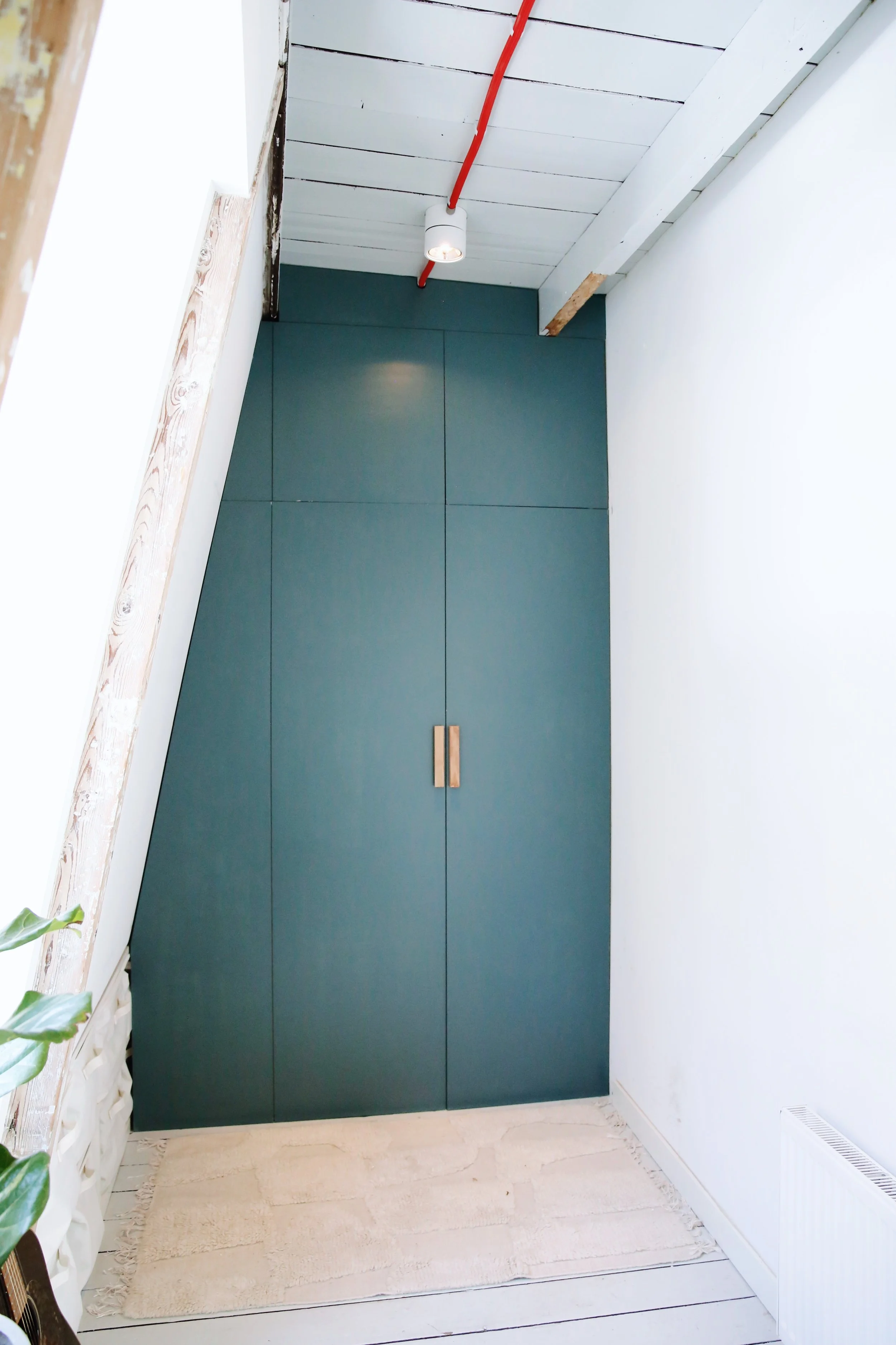 Groene kast in een witte kamer met houten vloer en een tapijt, plafond met witte houten planken, rode leiding met lamp, en een radiator.