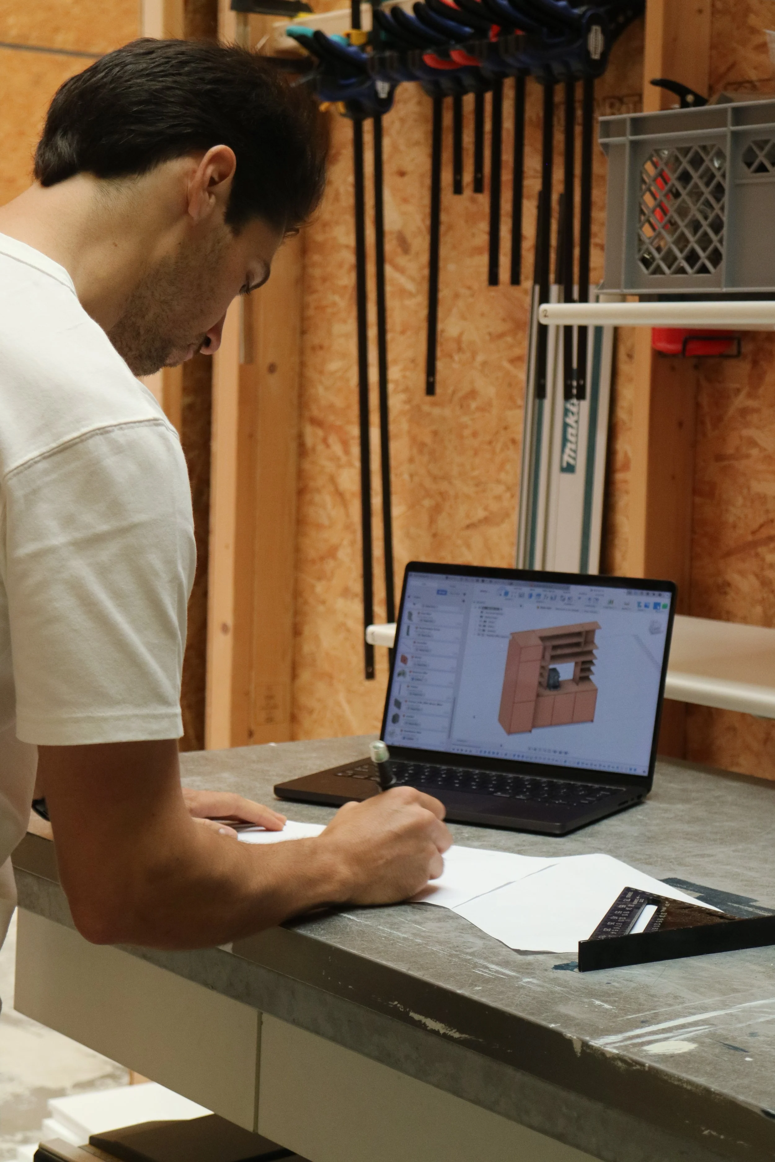 Man die een meubelontwerp tekent op papier in een werkplaats, met laptop en gereedschap op de tafel.