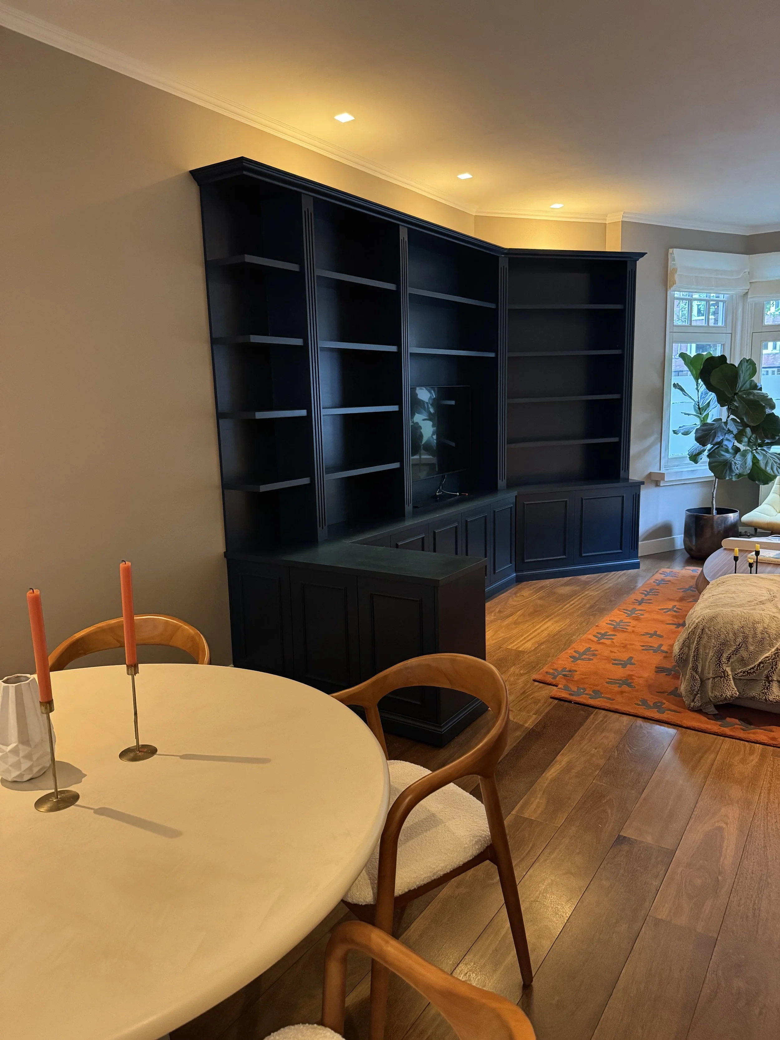 Een zwart ingebouwde boekenwand met open planken en een televisie in een woonkamer, naast een raam met wit raamwerk en groene planten, en een eethoek met een witte tafel en houten stoelen met beige kussens.