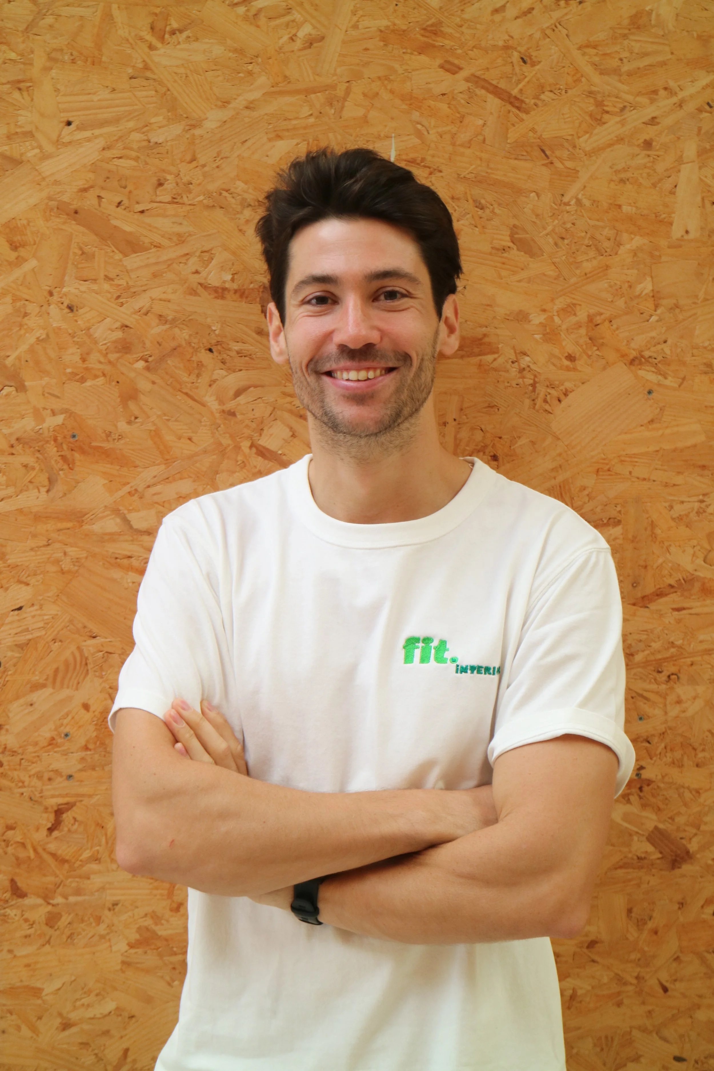 Een jonge man met donkere haar en een brede glimlach draagt een witte t-shirt met groene tekst 'fit interior'. Achtergrond van houten panelen.